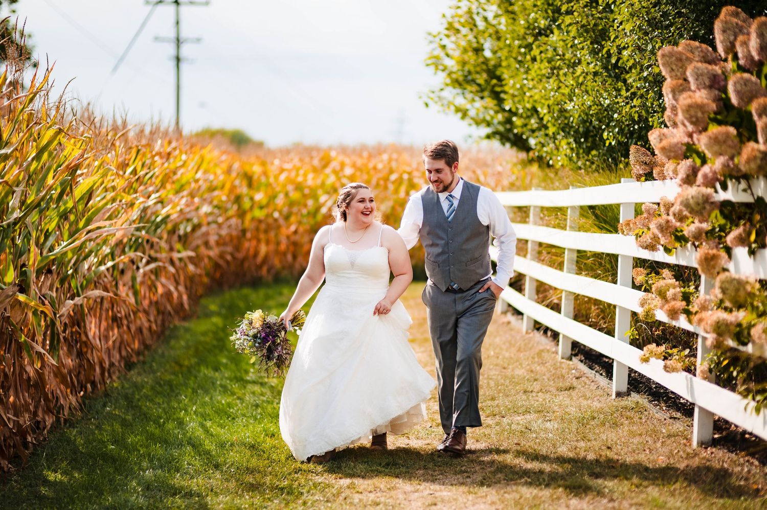 Couple walks along white fence with golden cornfield in autumn afternoon light.