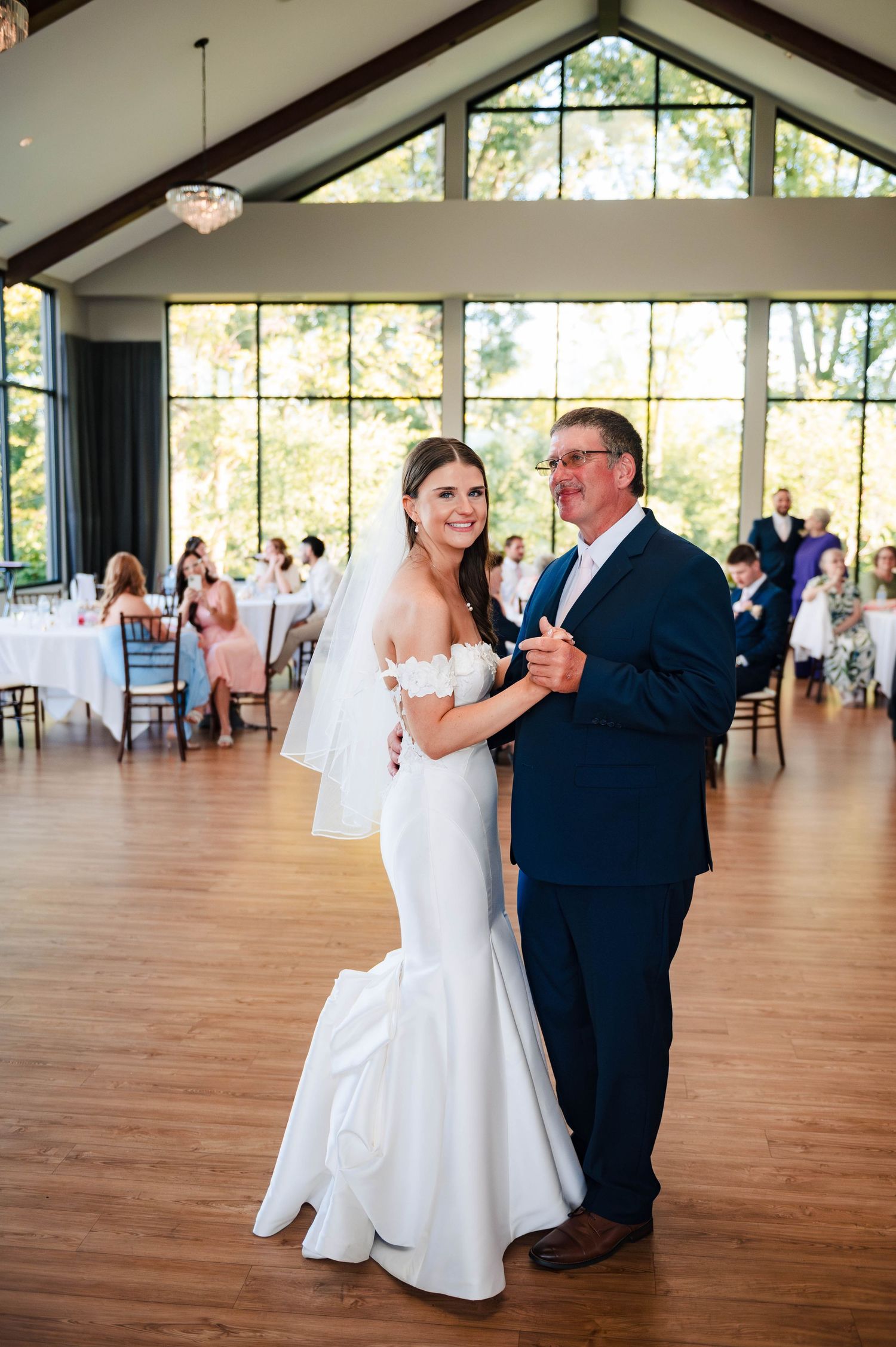 A wedding couple dances in an elegant reception venue with floor-to-ceiling windows.