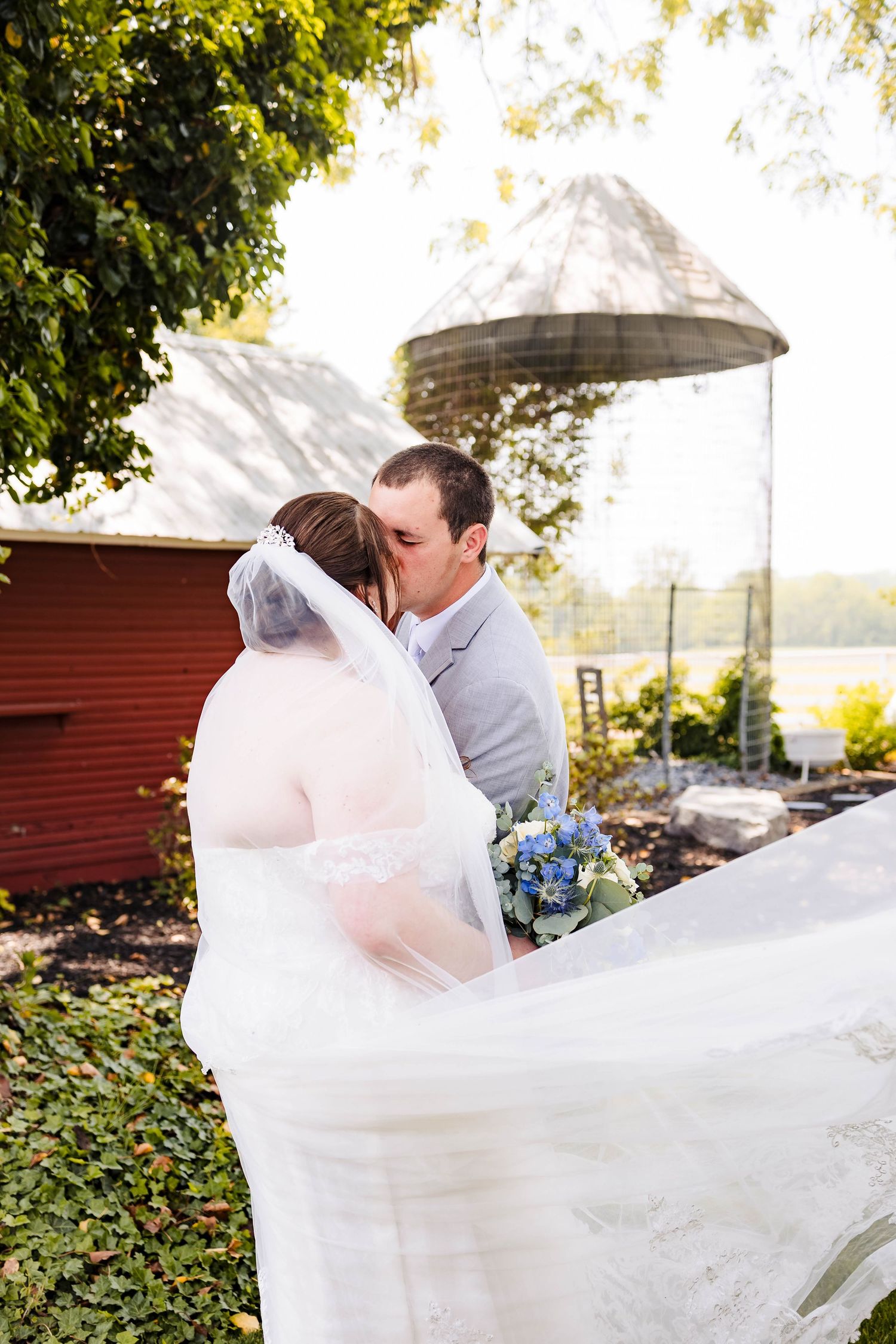 A romantic wedding couple embraces as the bride's veil flows dramatically in the wind at an outdoor venue.