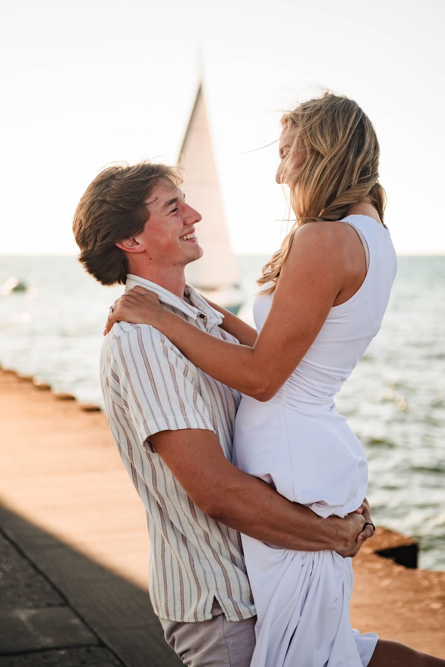 Playful couple embraces by red lighthouse with water in background.