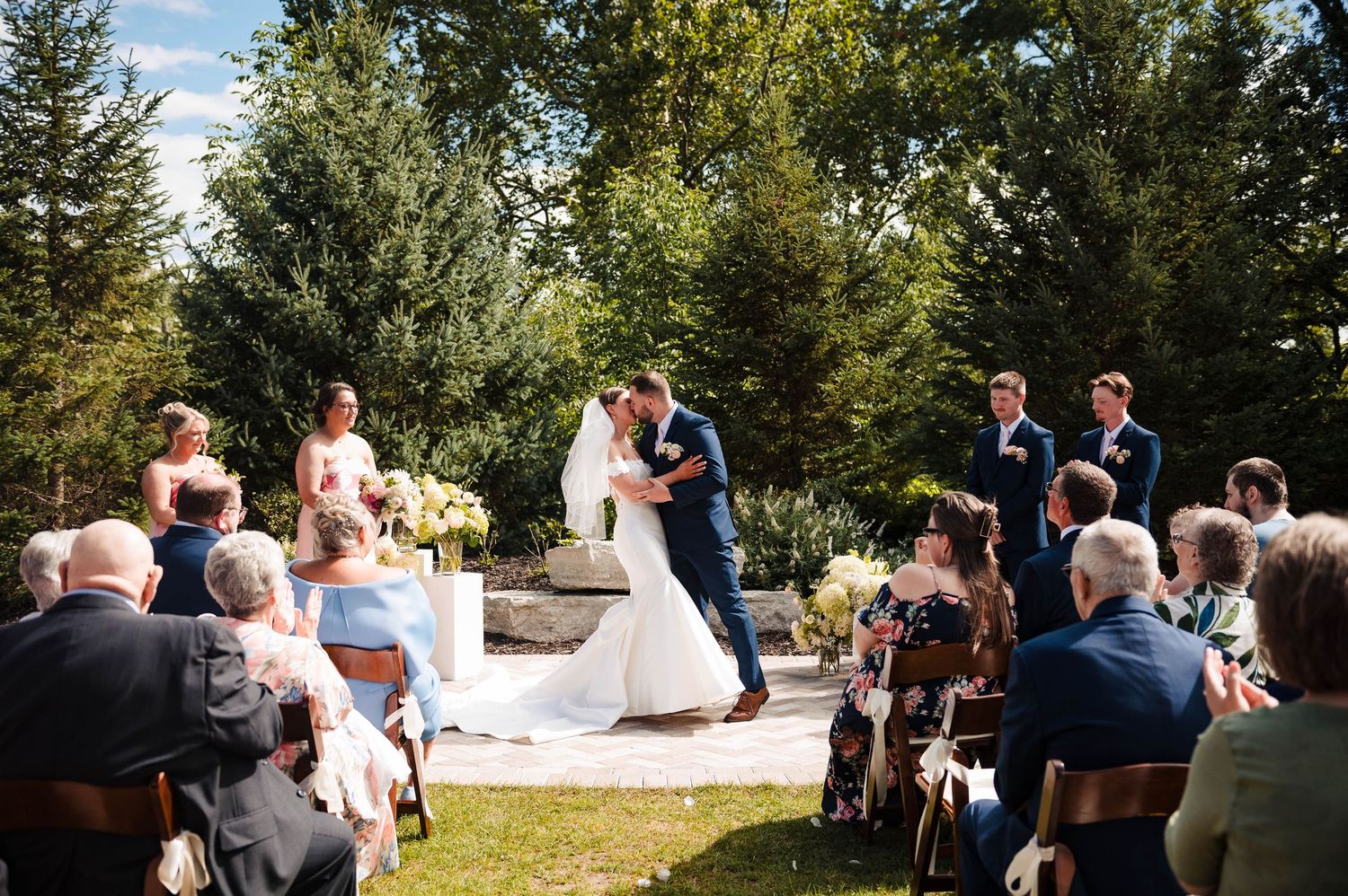 Outdoor wedding ceremony with guests seated among trees and greenery.