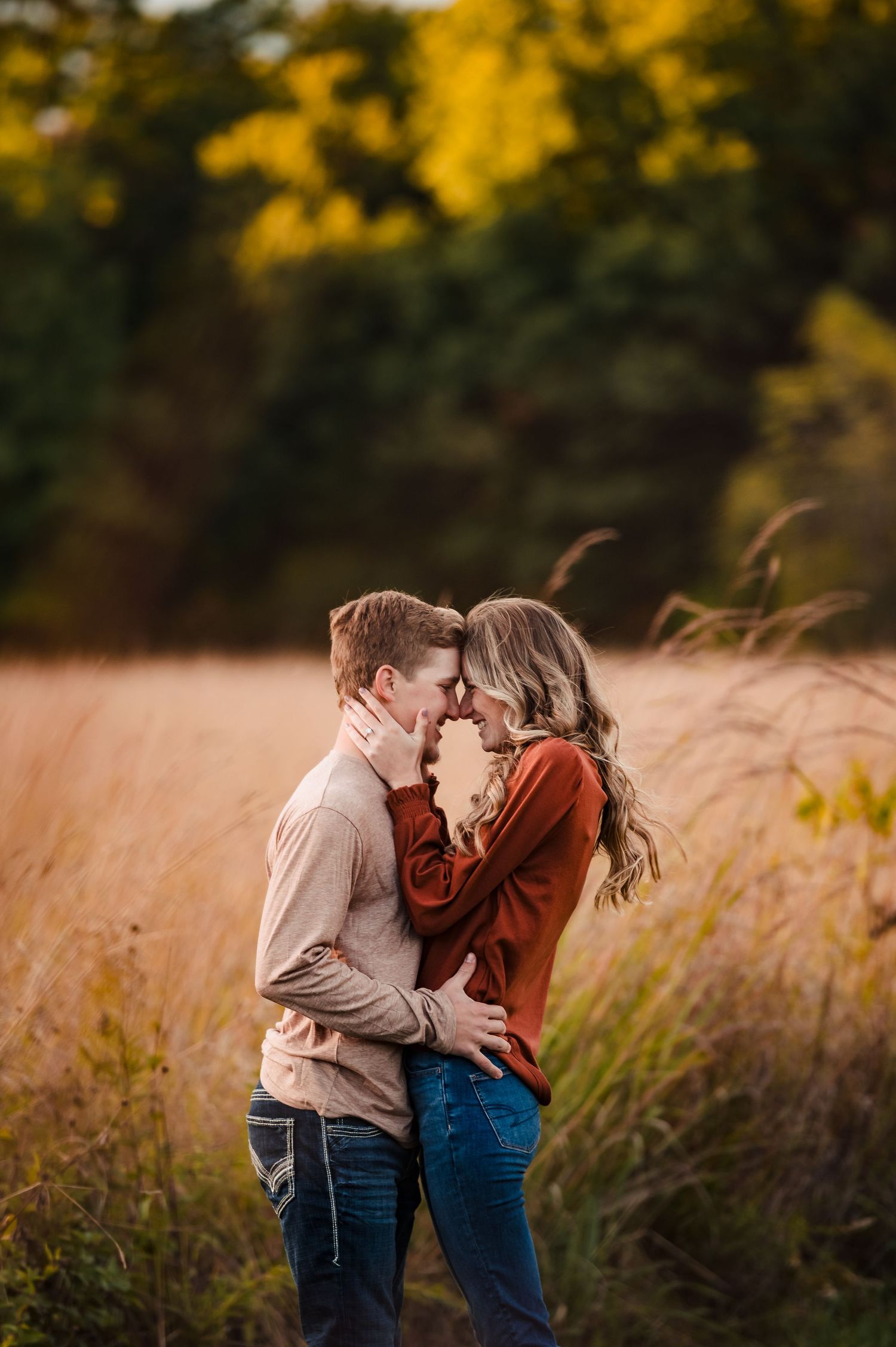 A romantic couple embraces in a golden autumn field during sunset with tall grass and trees in the background.