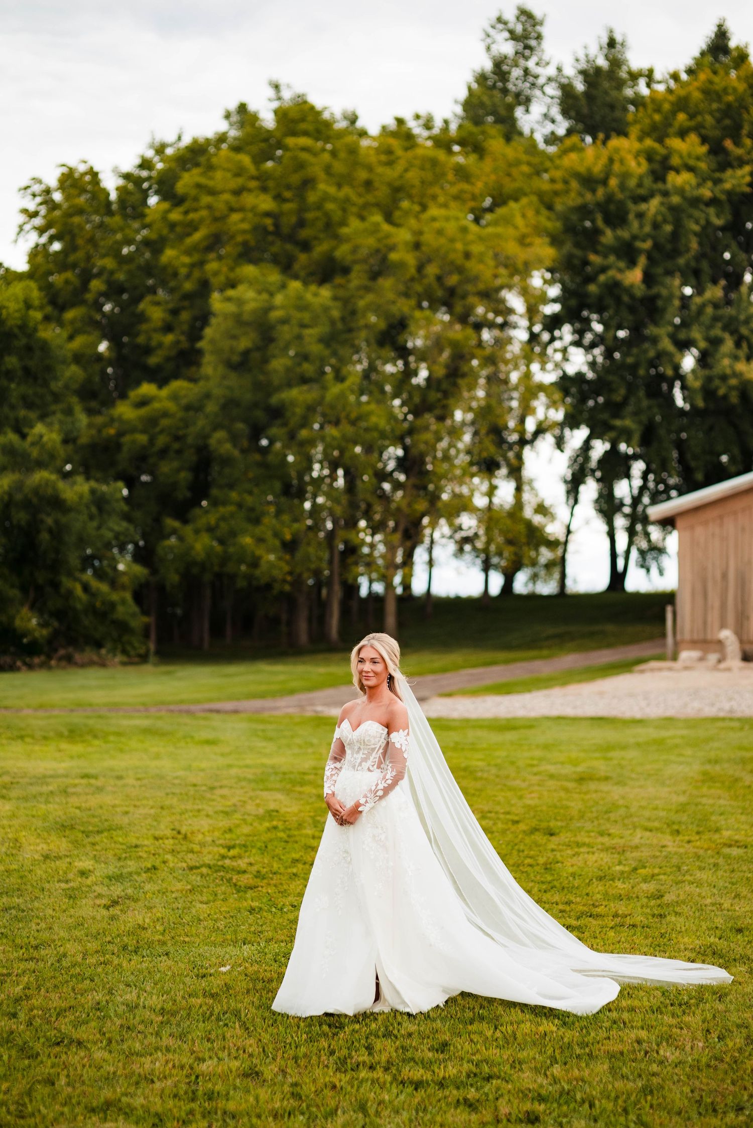 Bride in flowing white gown and veil poses on grass lawn with rustic barn in background.