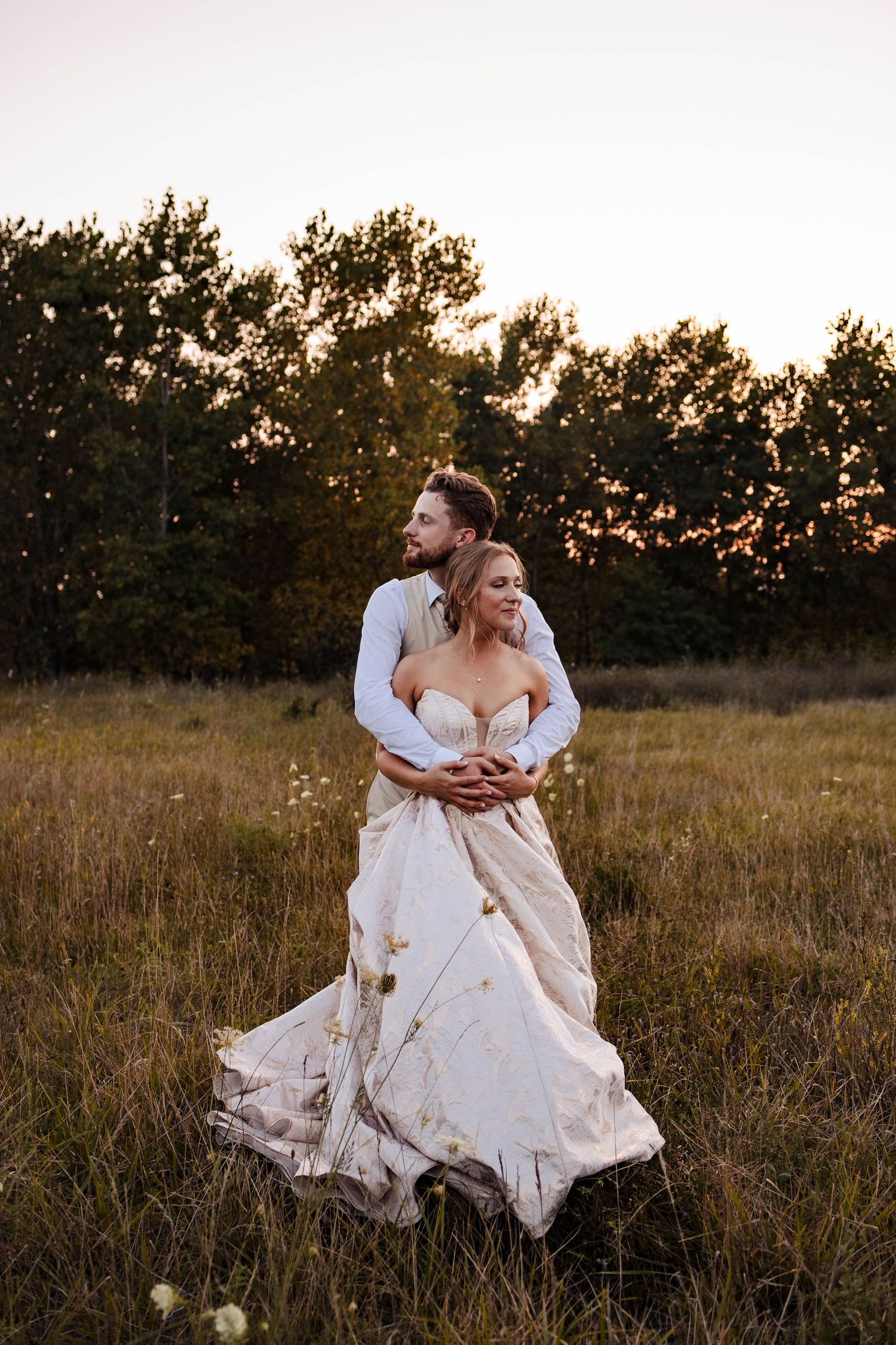 Romantic sunset portrait of newlyweds embracing in a rustic field with warm lighting.