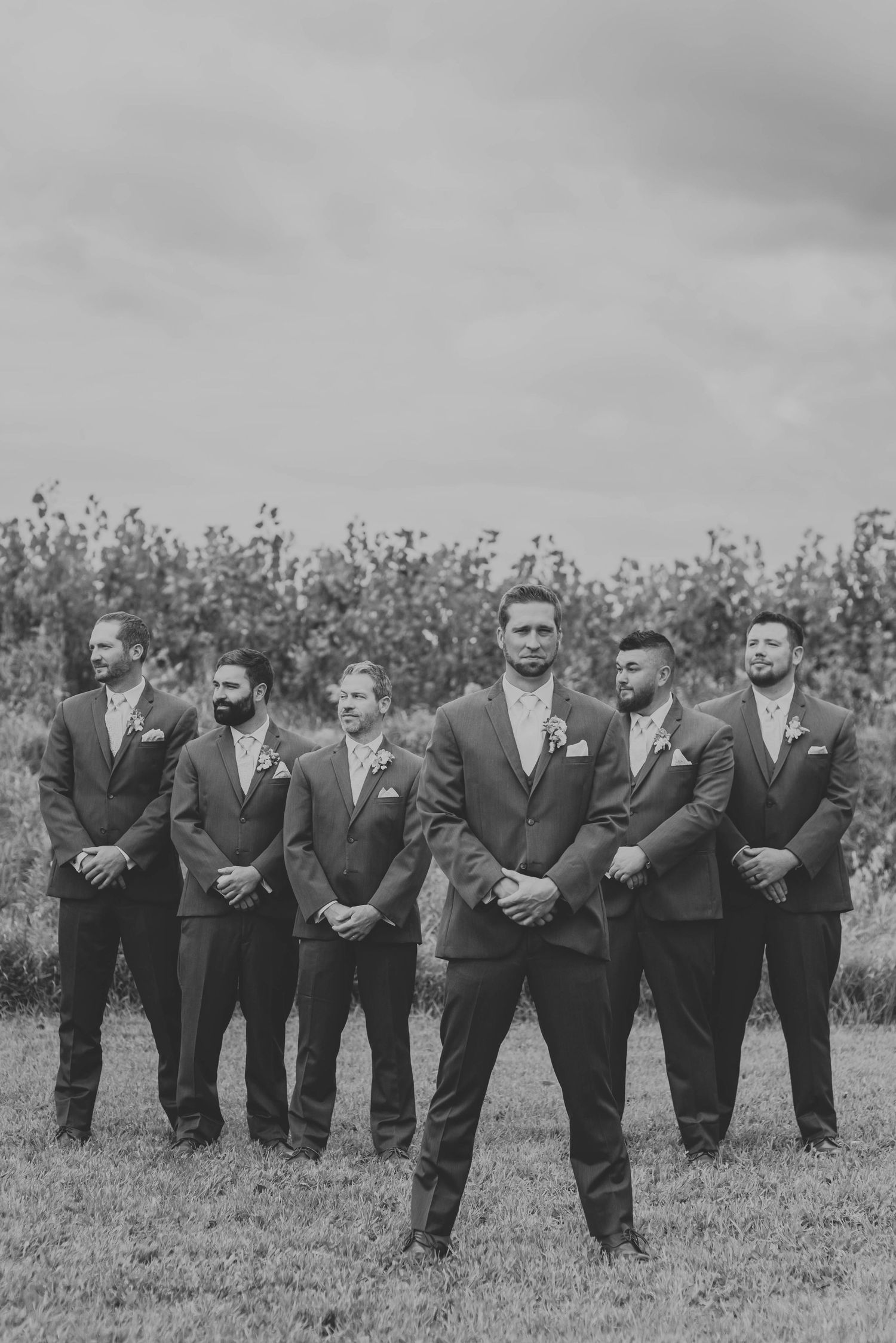 Black and white portrait of groomsmen in dark suits standing in formation against natural backdrop.