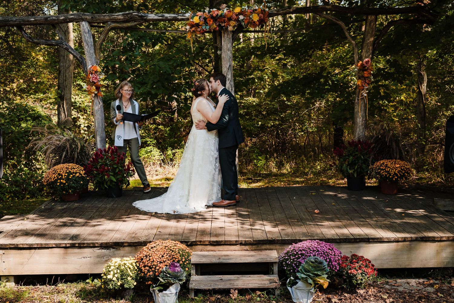 Wedding ceremony at a rustic outdoor venue with wooden stage decorated with autumn flowers.