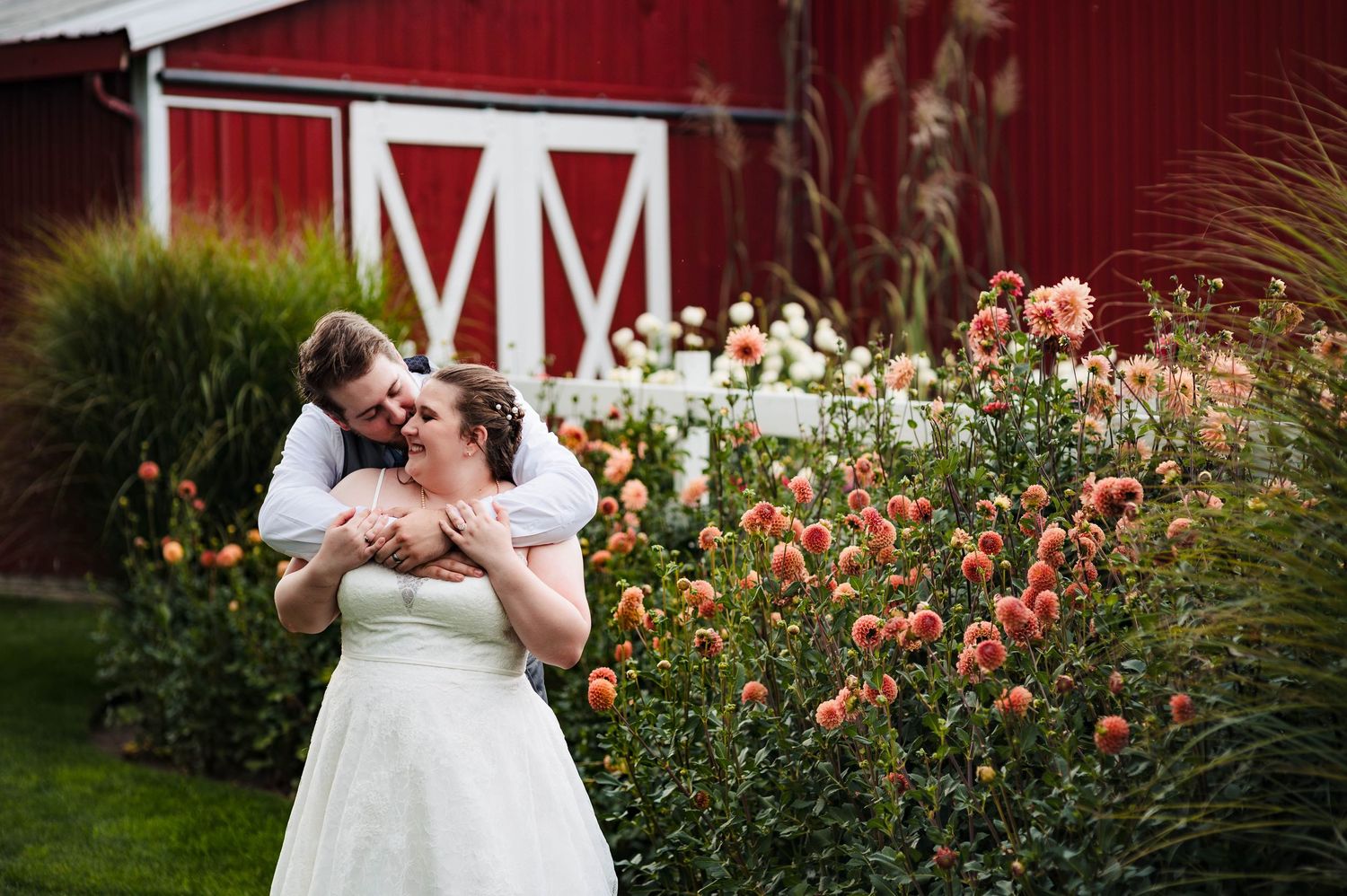 A wedding couple shares a romantic moment in front of a red barn with colorful autumn flowers.