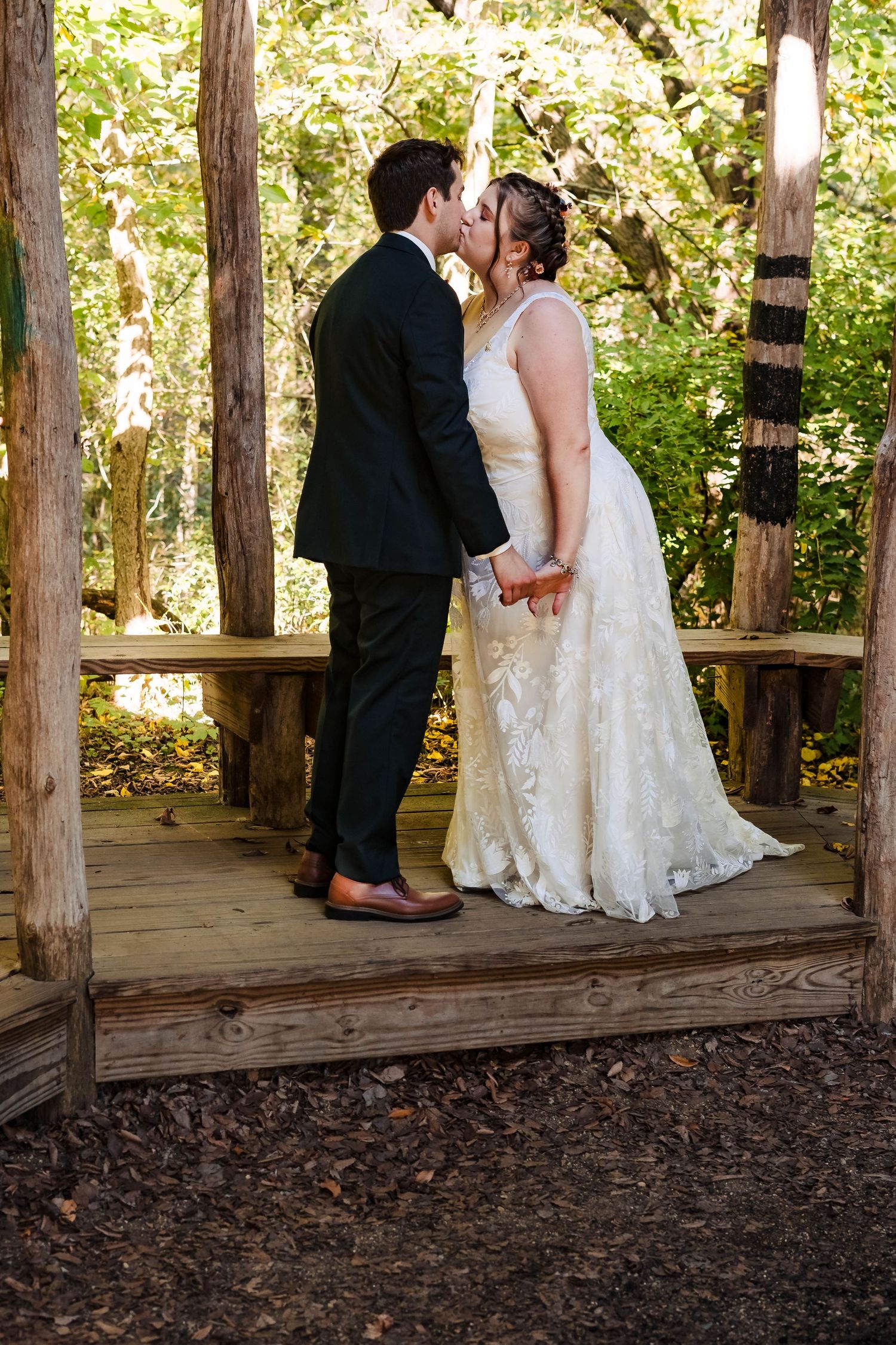 Wedding couple embraces on wooden deck surrounded by tall trees.
