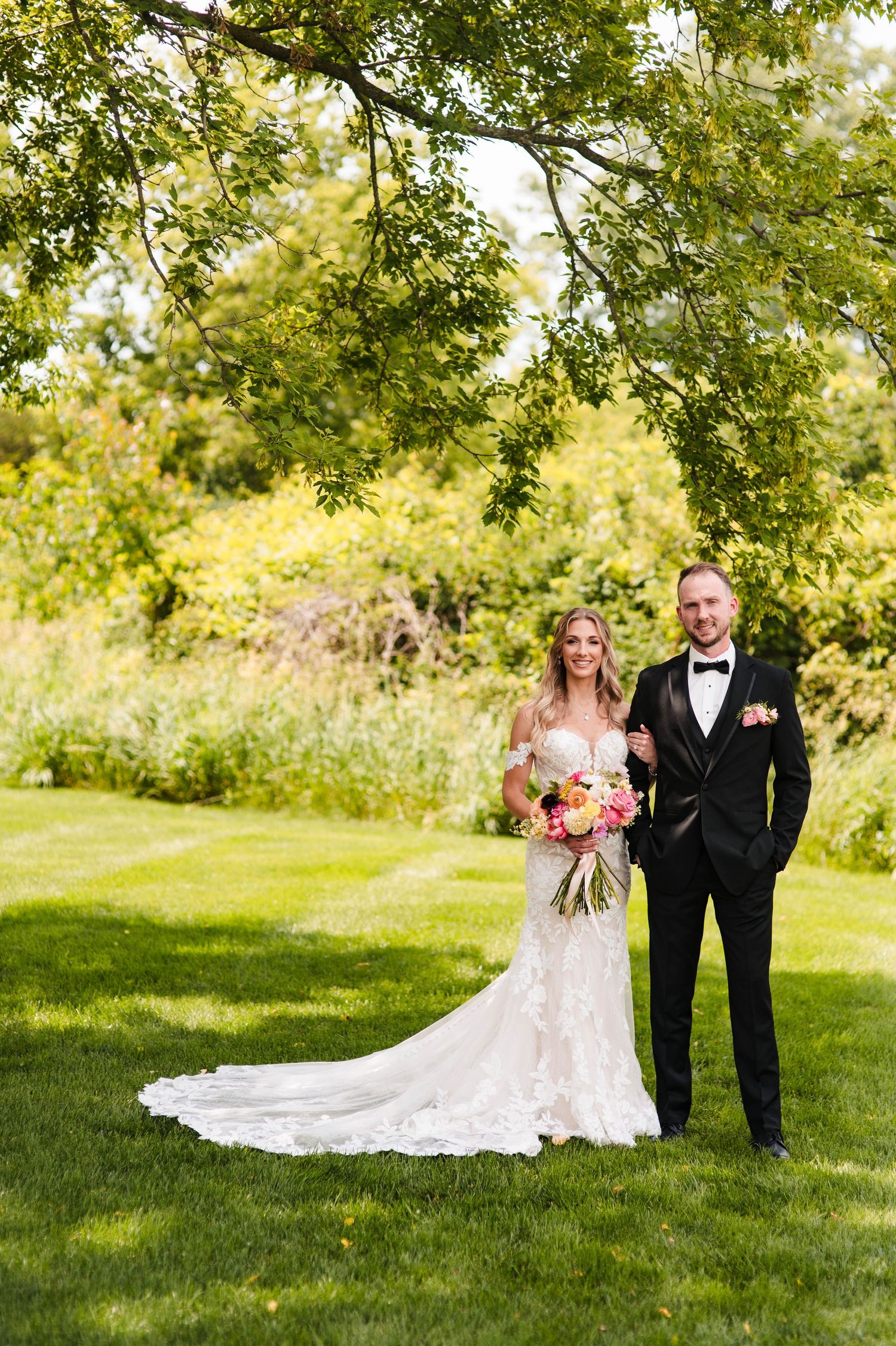 A couple in wedding attire walks together on a lush green lawn under sunlit trees during an outdoor summer celebration.
