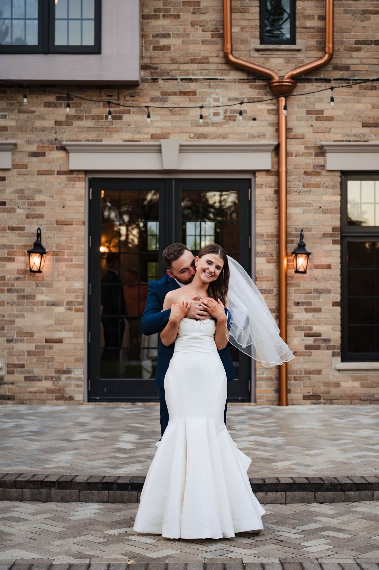 A romantic moment captured outside a brick building with copper accents and lanterns.