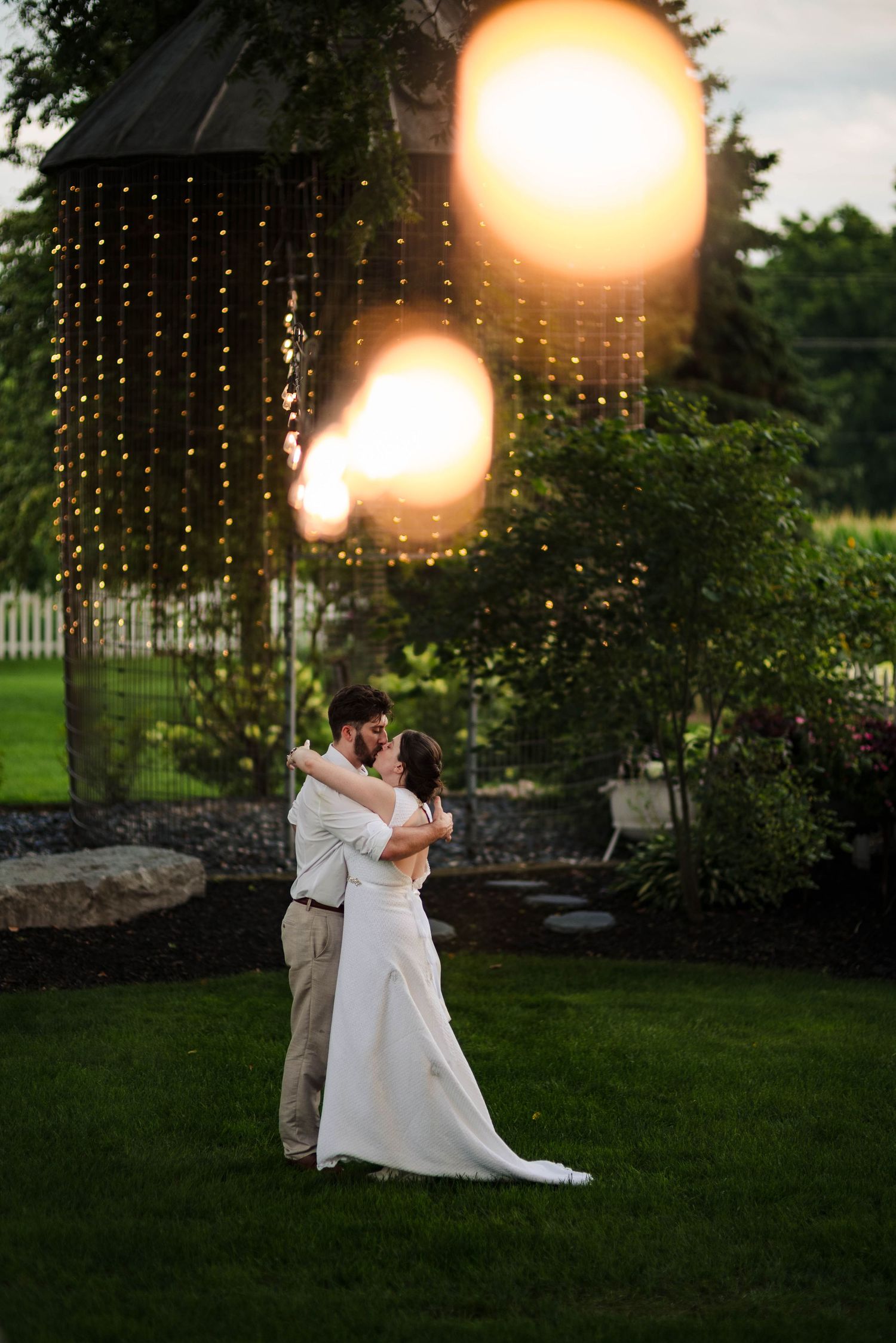 A romantic couple dances on a lawn at sunset with string lights glowing overhead.