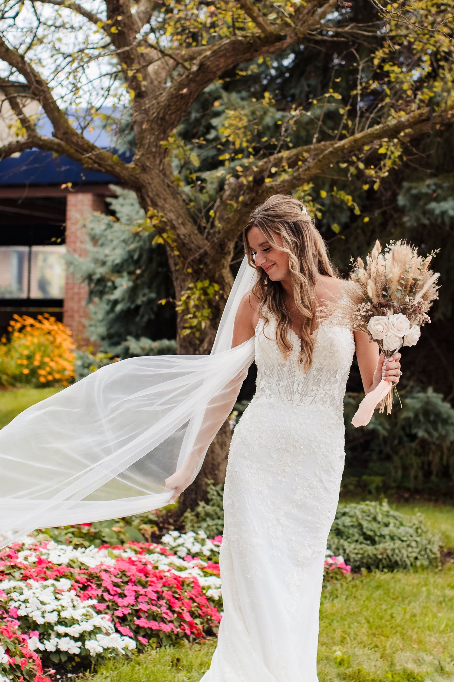 A bride in a flowing white dress twirls gracefully in a garden with pink flowers and autumn trees in the background.