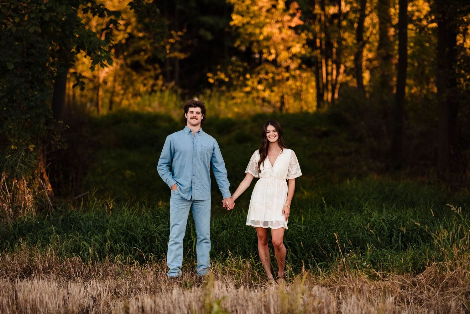 Couple walking through autumn field during golden hour with trees in background.