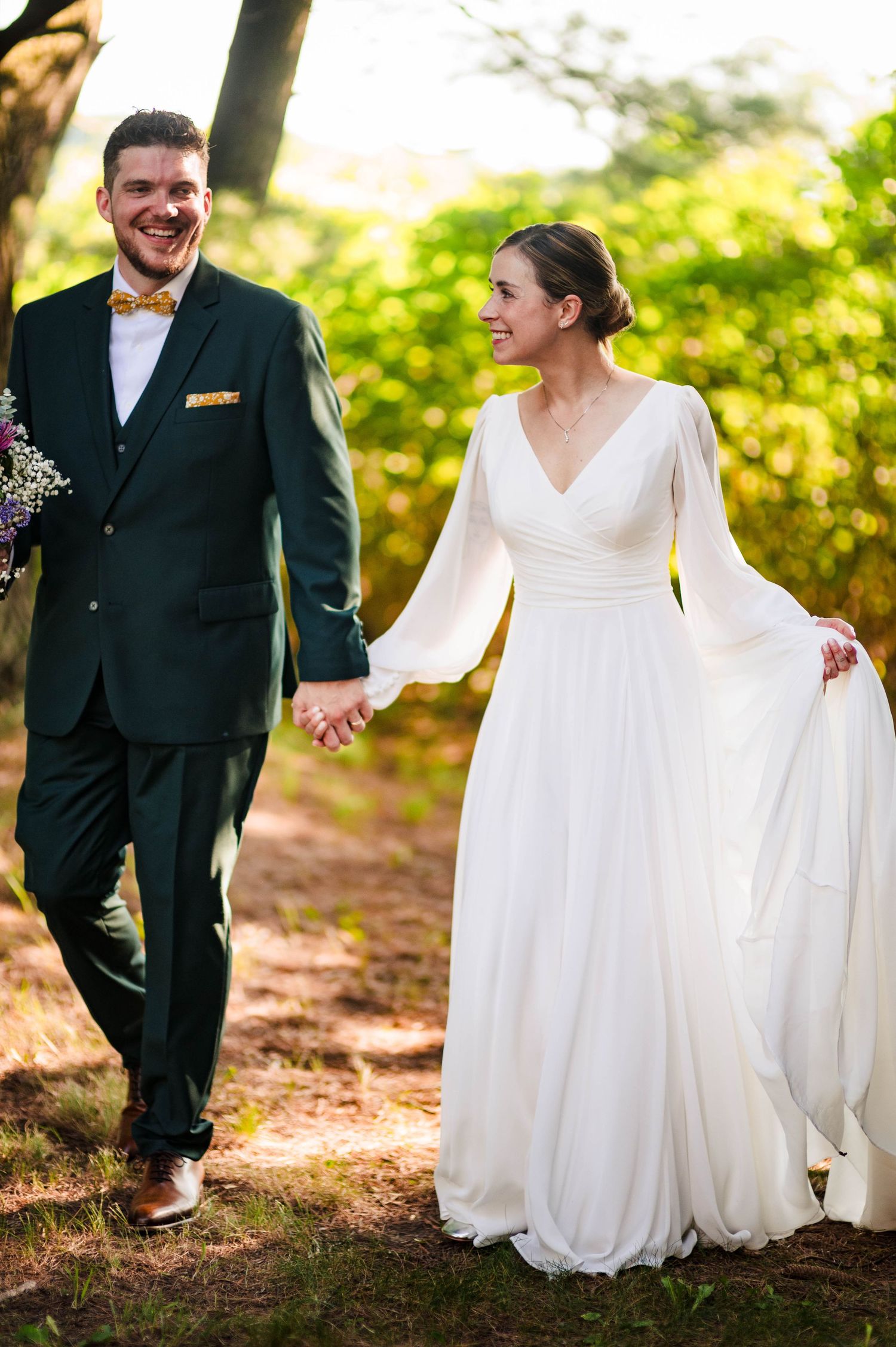 Bride in flowing white dress walks through garden path with groom in dark suit.