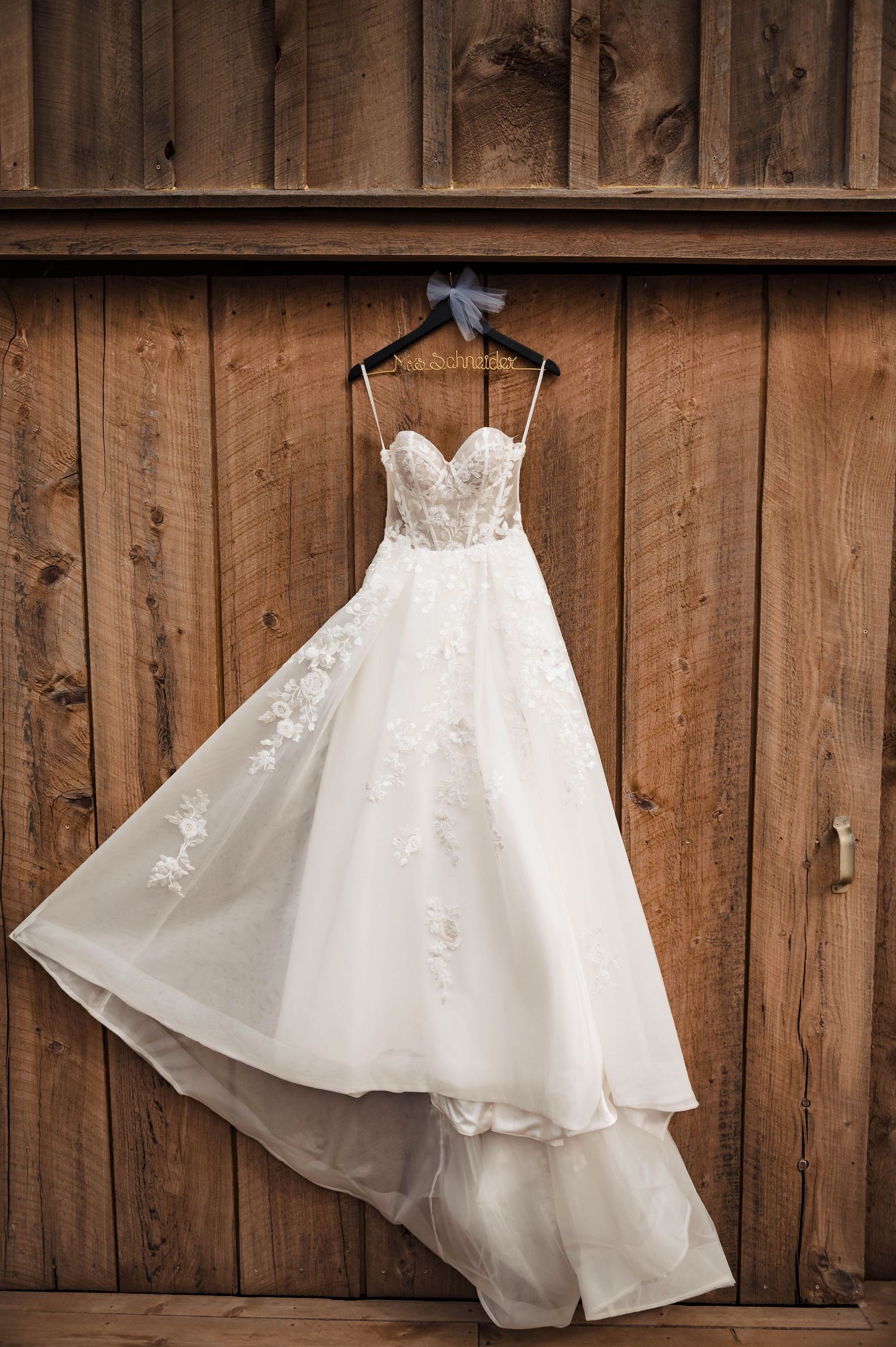 An elegant white wedding dress with lace details hangs on a rustic wooden wall.