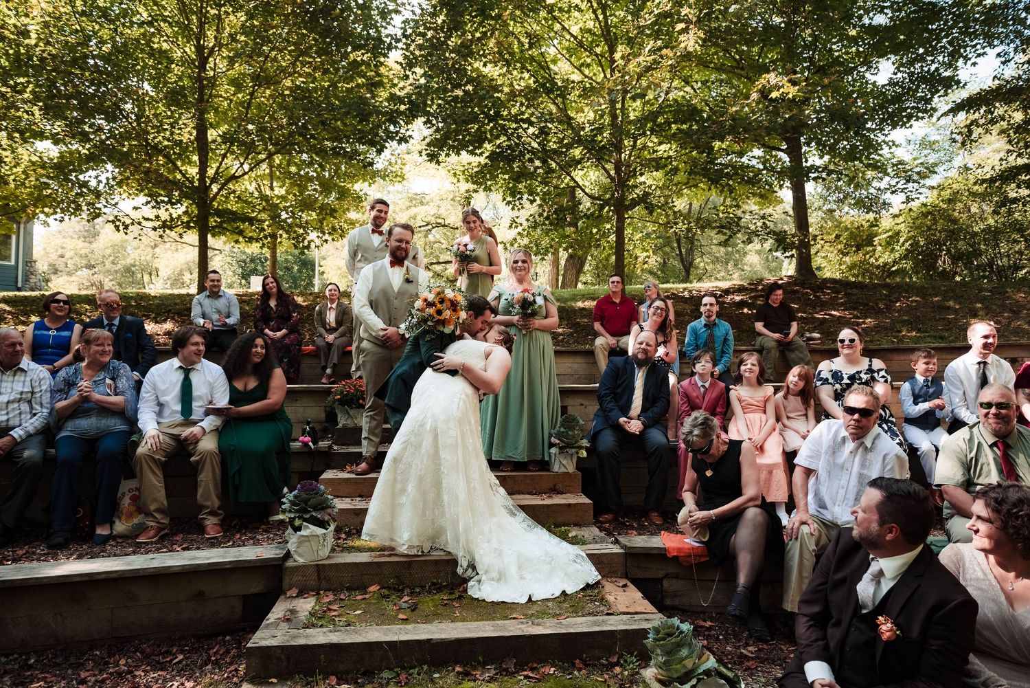 Wedding ceremony guests gather outdoors under green trees during a sunny afternoon celebration.