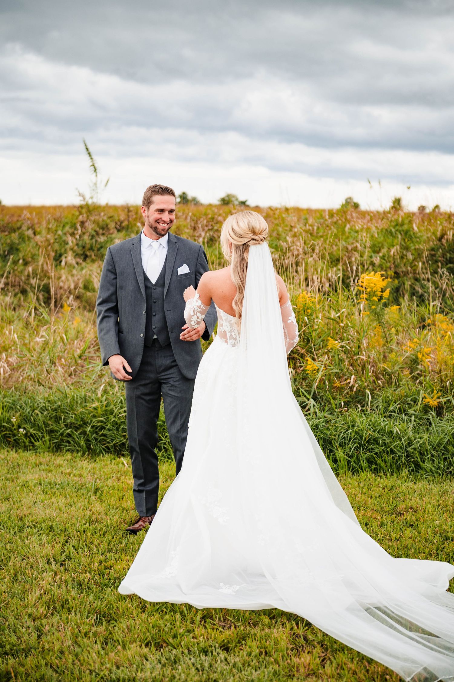 A bride in flowing white gown walks with groom through a meadow of wildflowers.