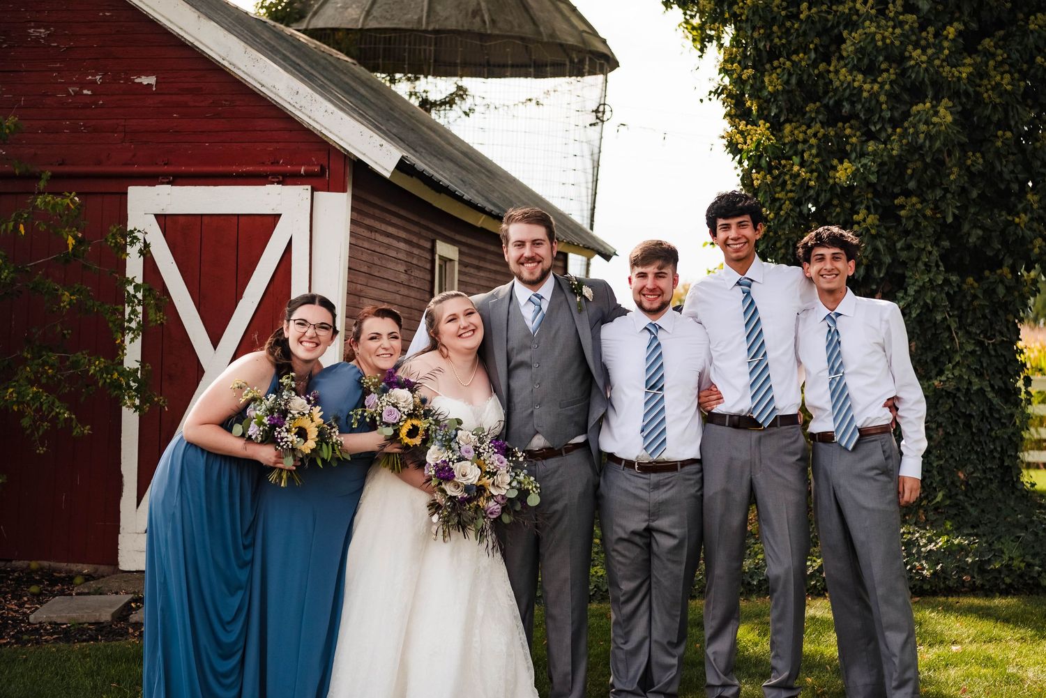 Wedding party poses in front of a red barn wearing blue and gray attire.