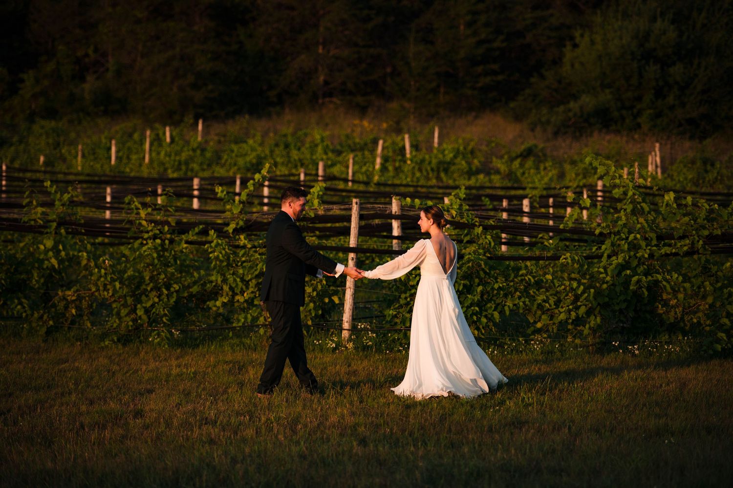 Silhouetted wedding couple walks along rustic fence line at sunset.