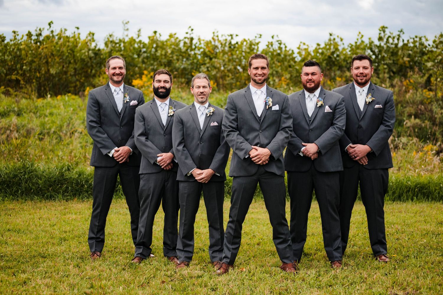 Groomsmen in matching grey and navy suits pose together outdoors against a natural landscape.