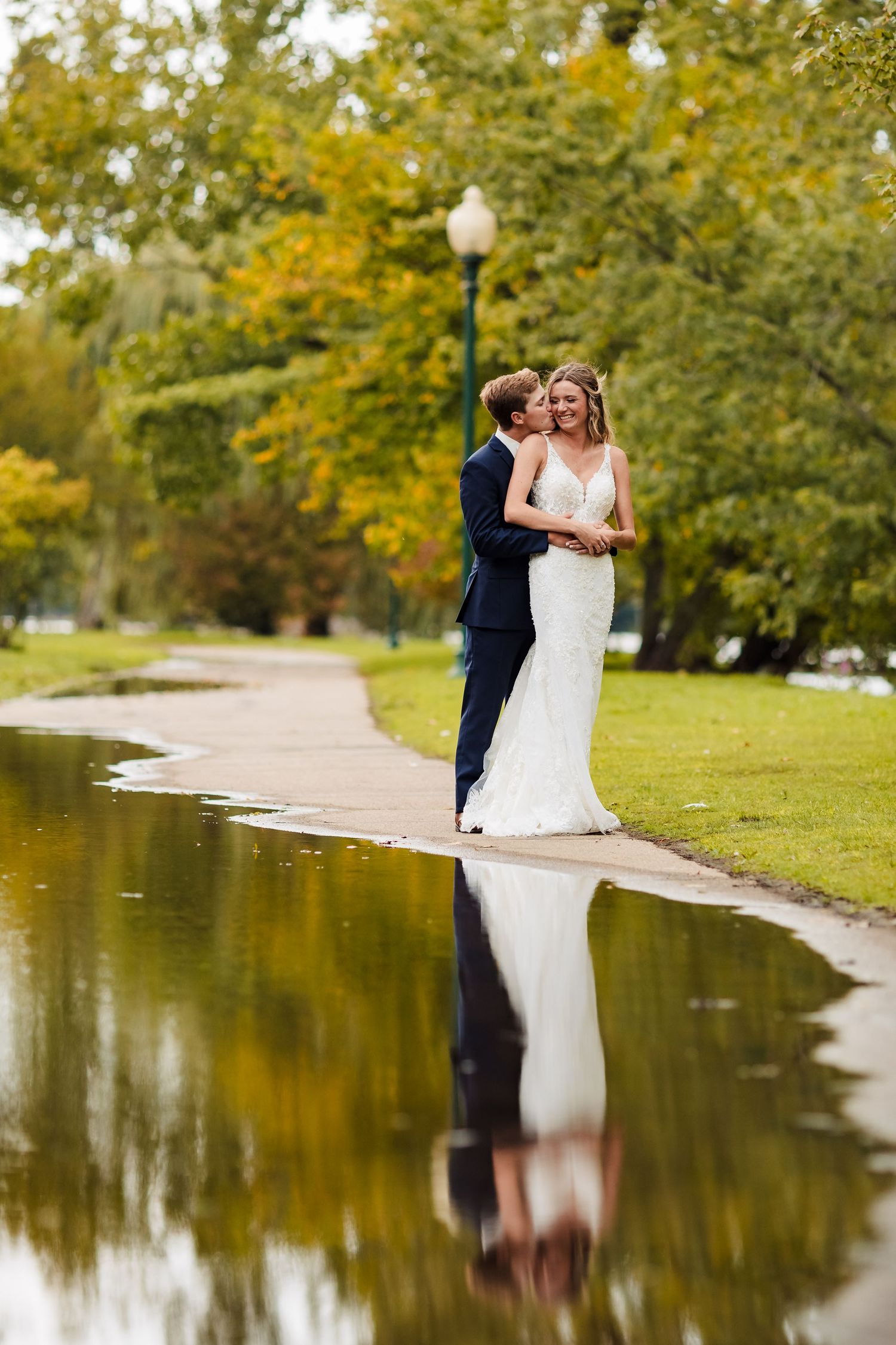 A romantic wedding couple embraces by a tranquil pond surrounded by autumn trees with golden leaves.