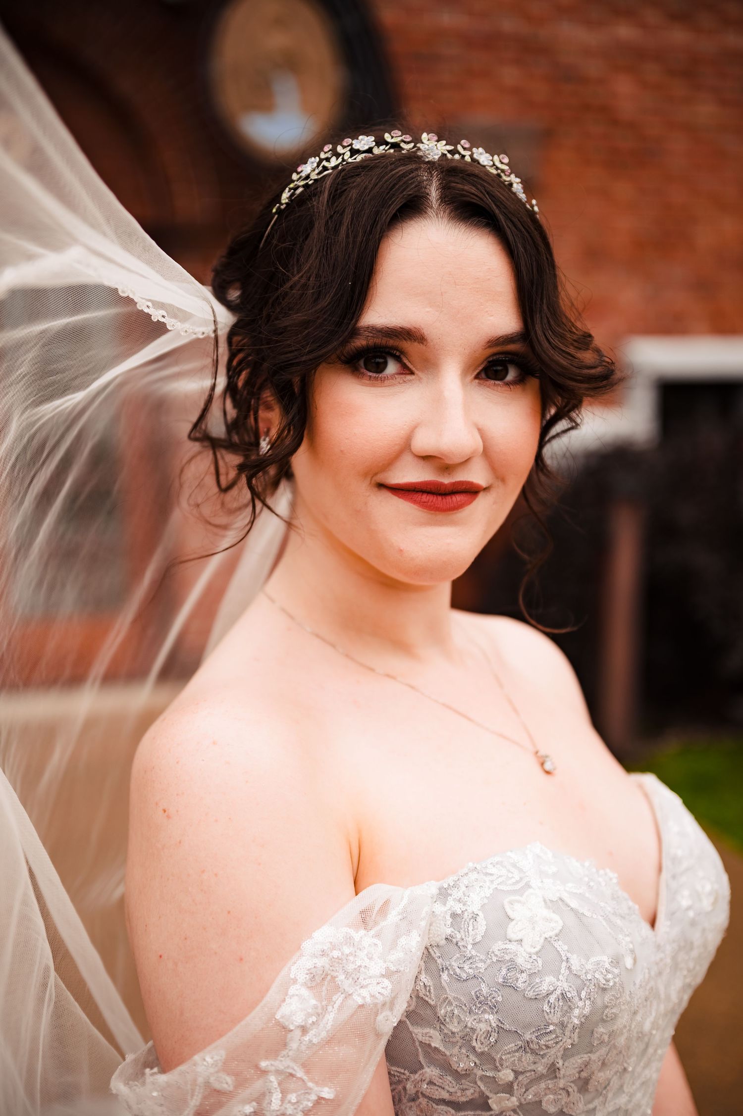 Bride wearing an elegant white lace wedding dress with sweetheart neckline and flowing veil poses against brick wall.