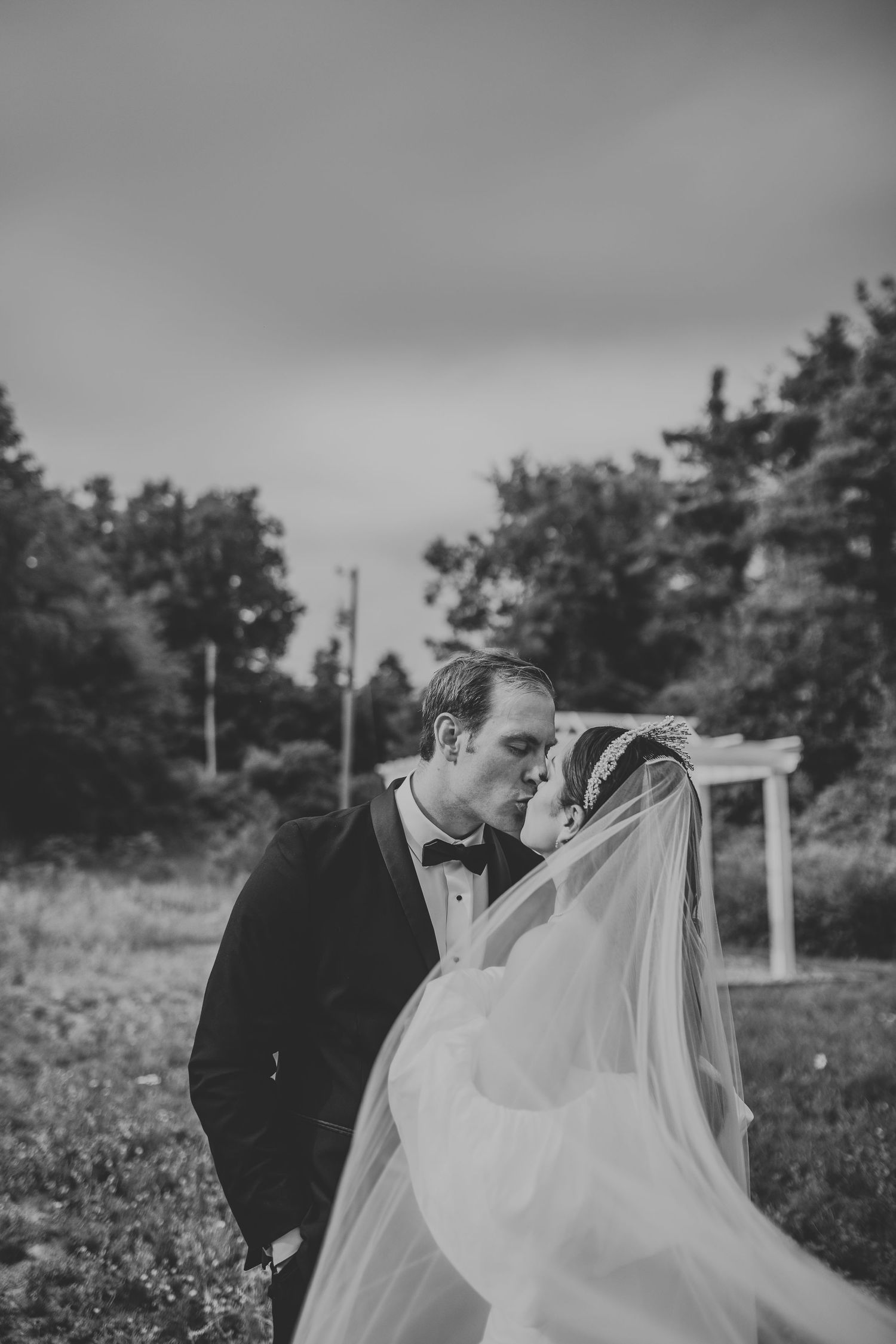 A romantic black and white wedding photo showing a tender kiss between newlyweds with a flowing veil in an outdoor setting.