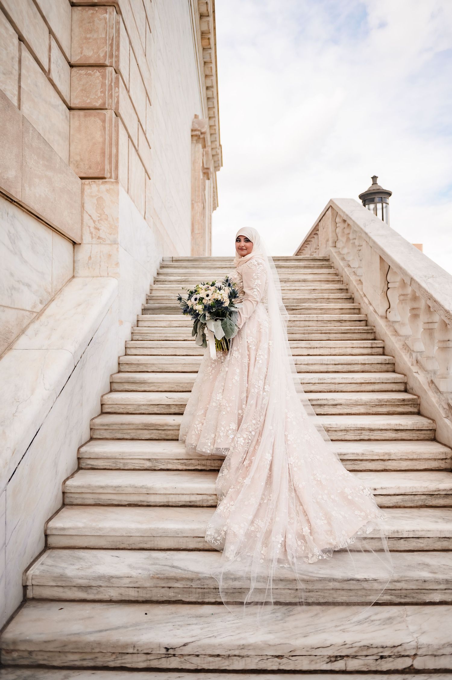 A bride in a flowing blush lace gown poses gracefully on elegant white marble stairs at a grand venue.