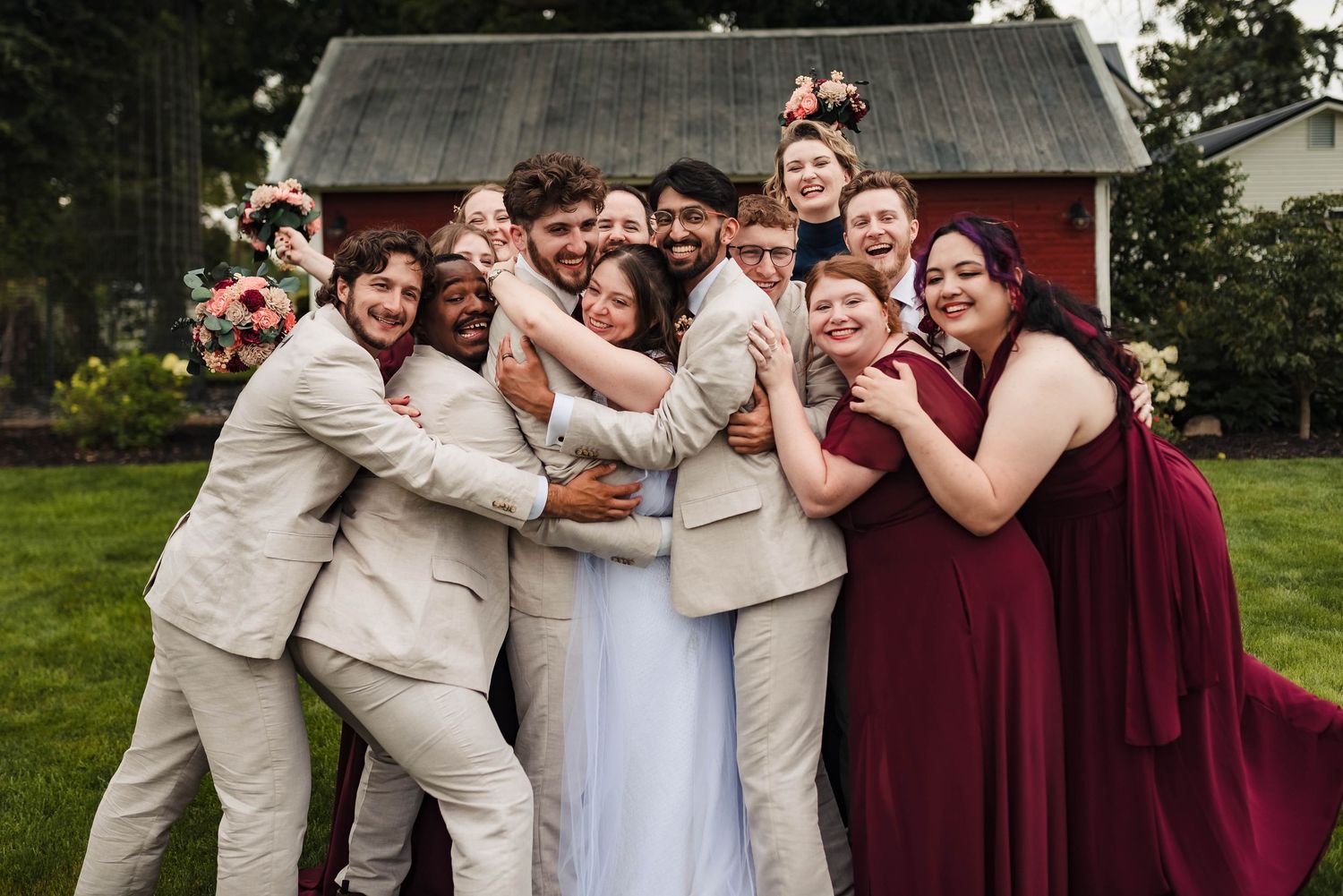 Wedding party in burgundy and beige attire posing playfully outside rustic barn.