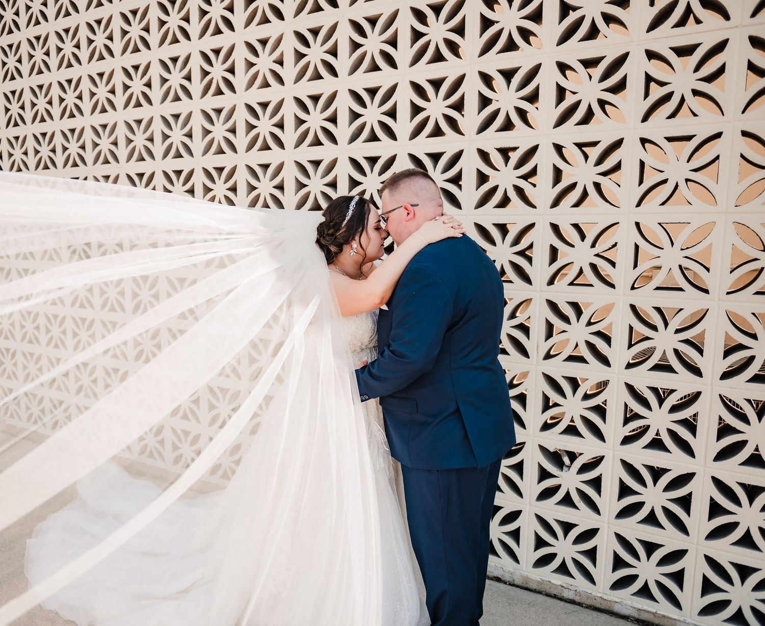 A couple in wedding attire embraces against a decorative geometric wall while a veil flows in the breeze.