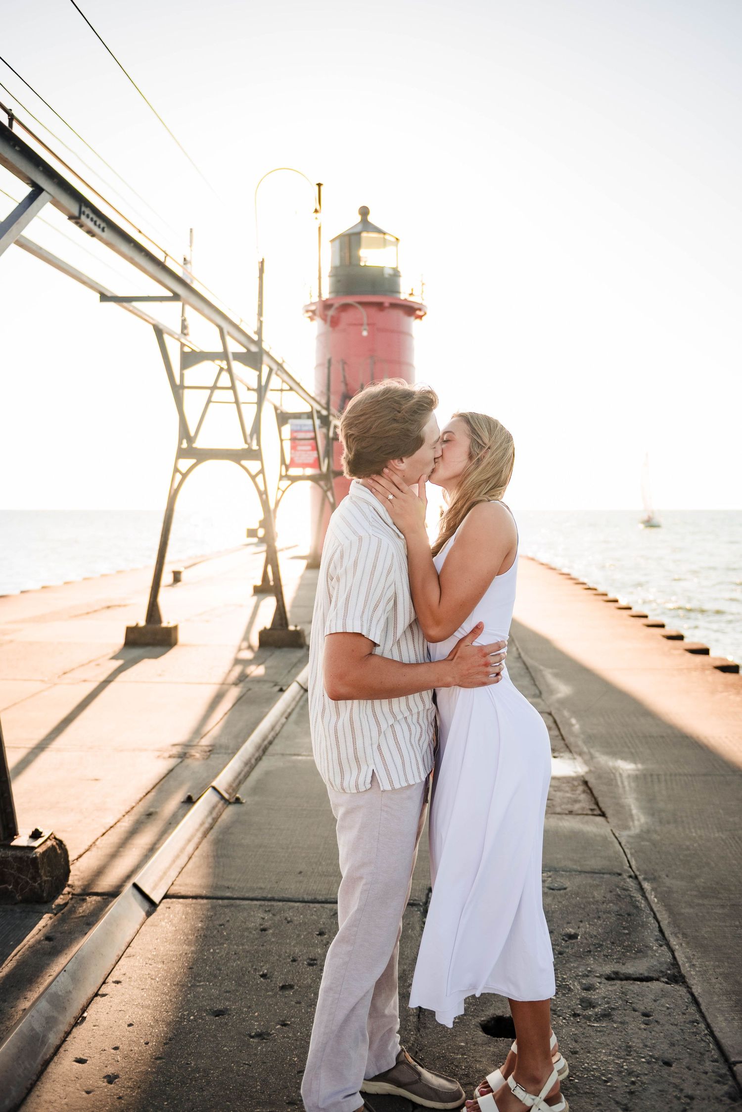 Couple sharing a romantic moment on a pier with lighthouse and sunset in the background.
