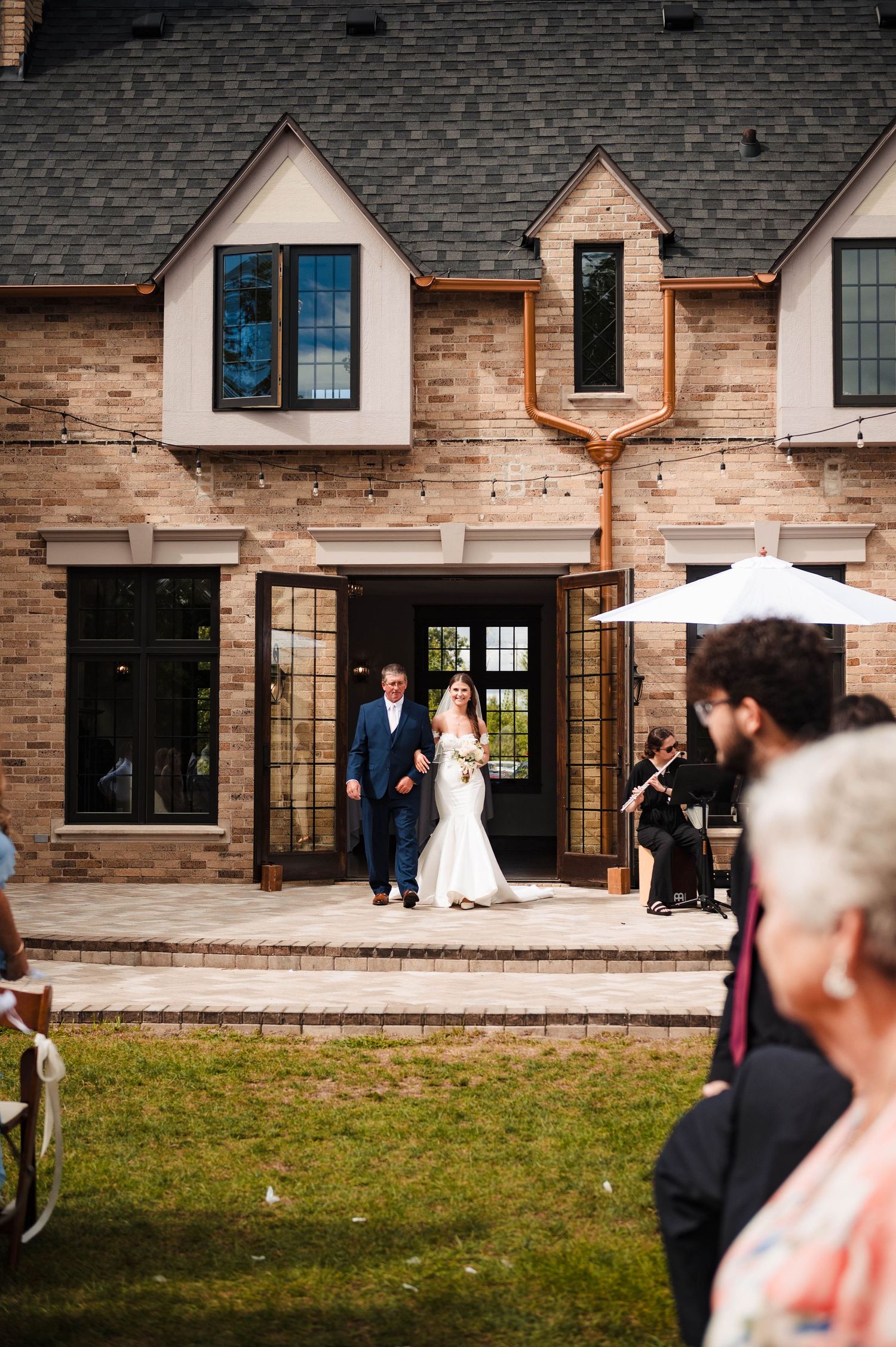 Newlyweds exit a beautiful brick venue with Tudor-style architecture as guests look on.