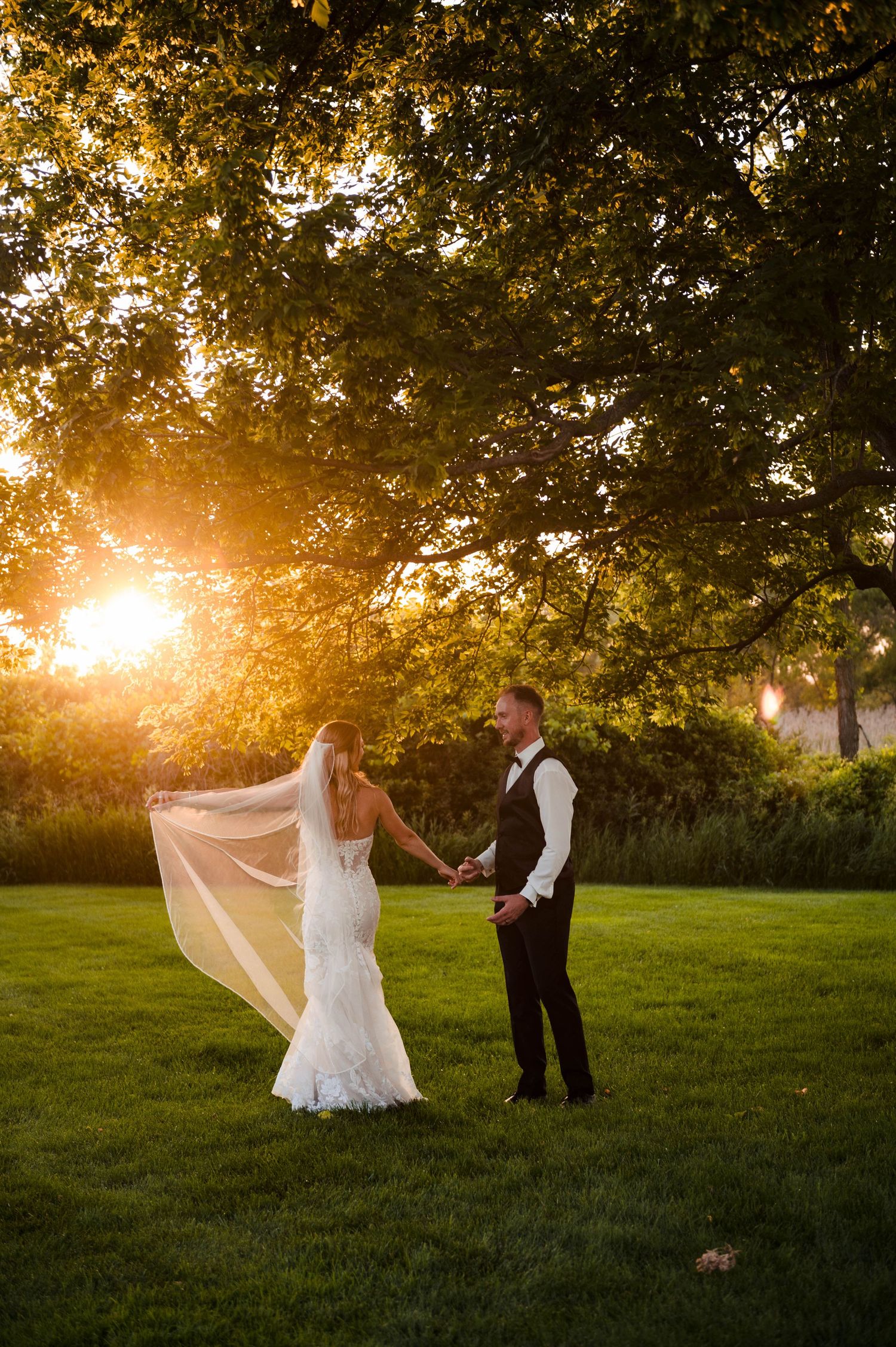 A couple dances together in a grassy field at sunset while a wedding veil flows in the golden light.