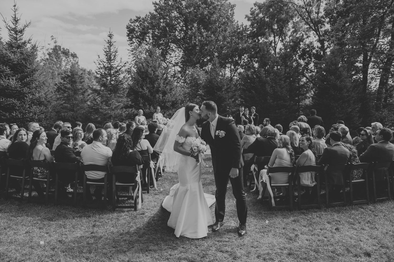 Black and white panorama of outdoor wedding ceremony with guests seated among trees.
