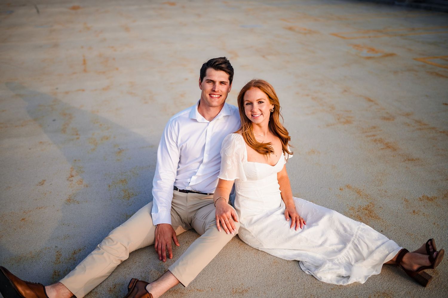 A couple in elegant attire sits together on a concrete surface during a romantic photoshoot.