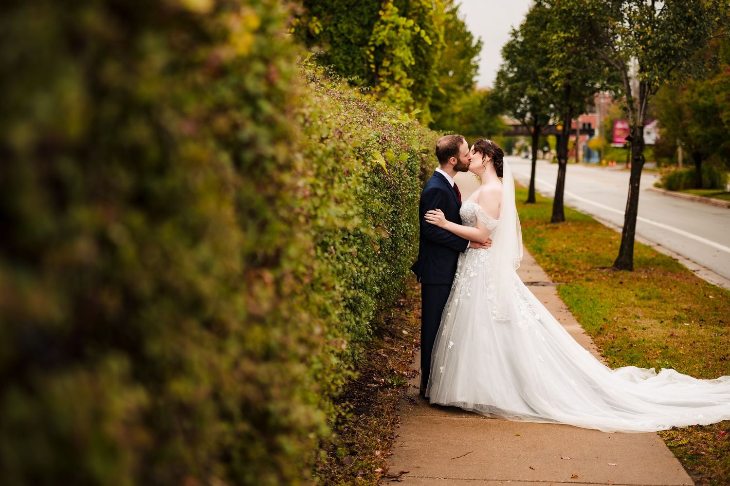 Couple in formal wedding attire embraces on a tree-lined autumn path during their romantic outdoor photoshoot.