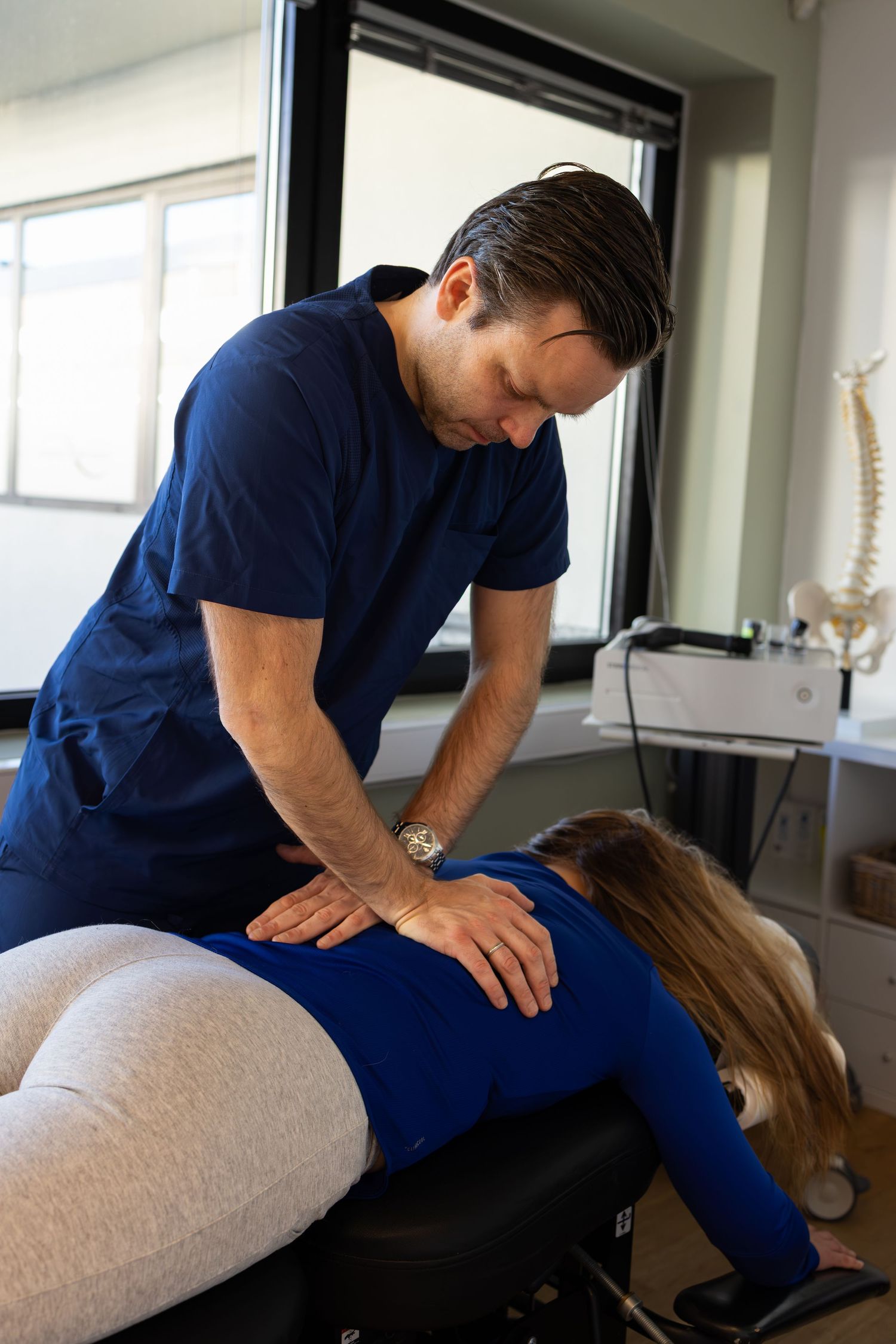 Chiropractor performs spinal adjustment therapy on patient lying on treatment table in medical office.