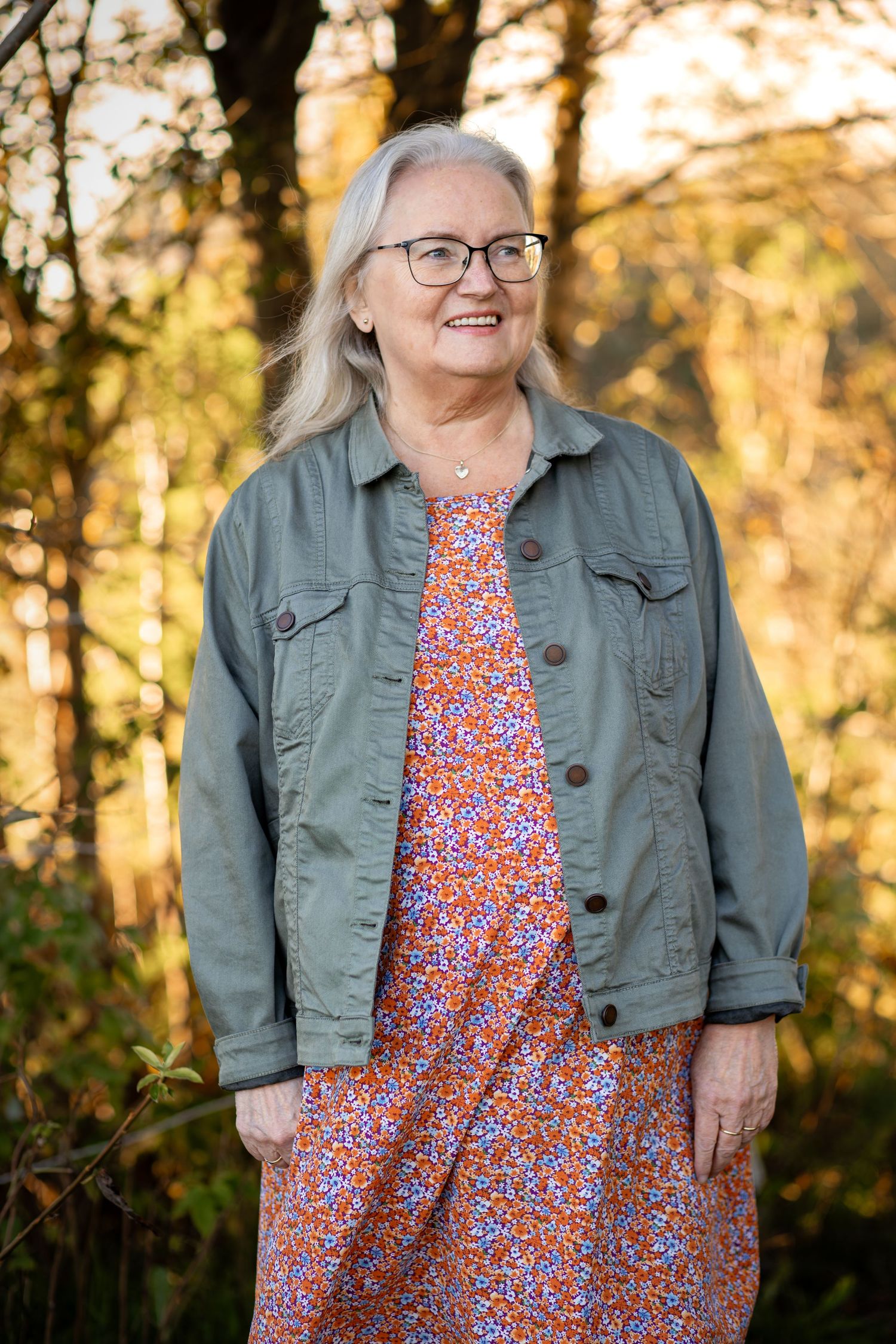 A person wearing glasses and a denim jacket over a floral dress stands in a sunlit autumn forest setting.