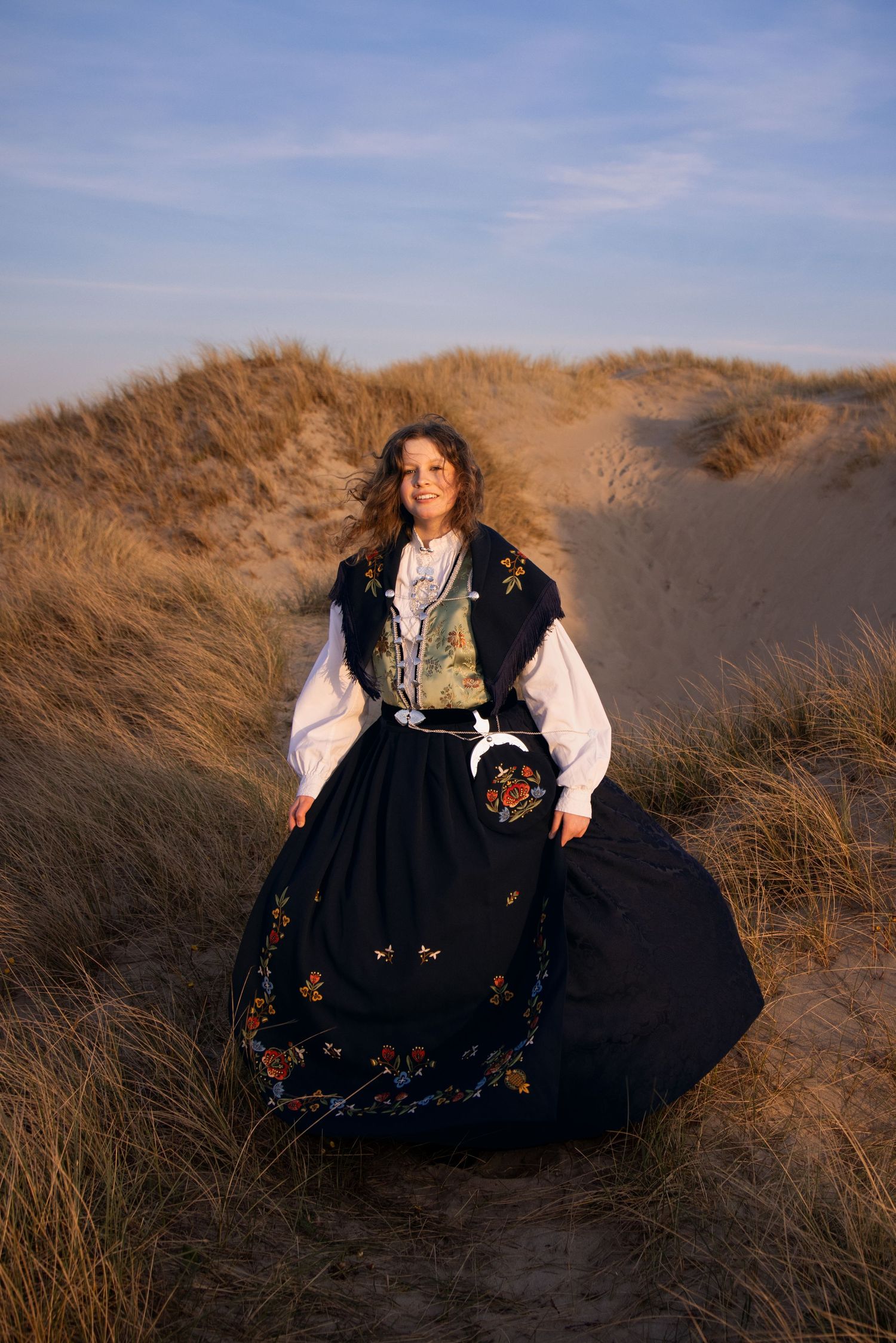 A person in traditional Norwegian folk dress stands among sand dunes at golden hour.
