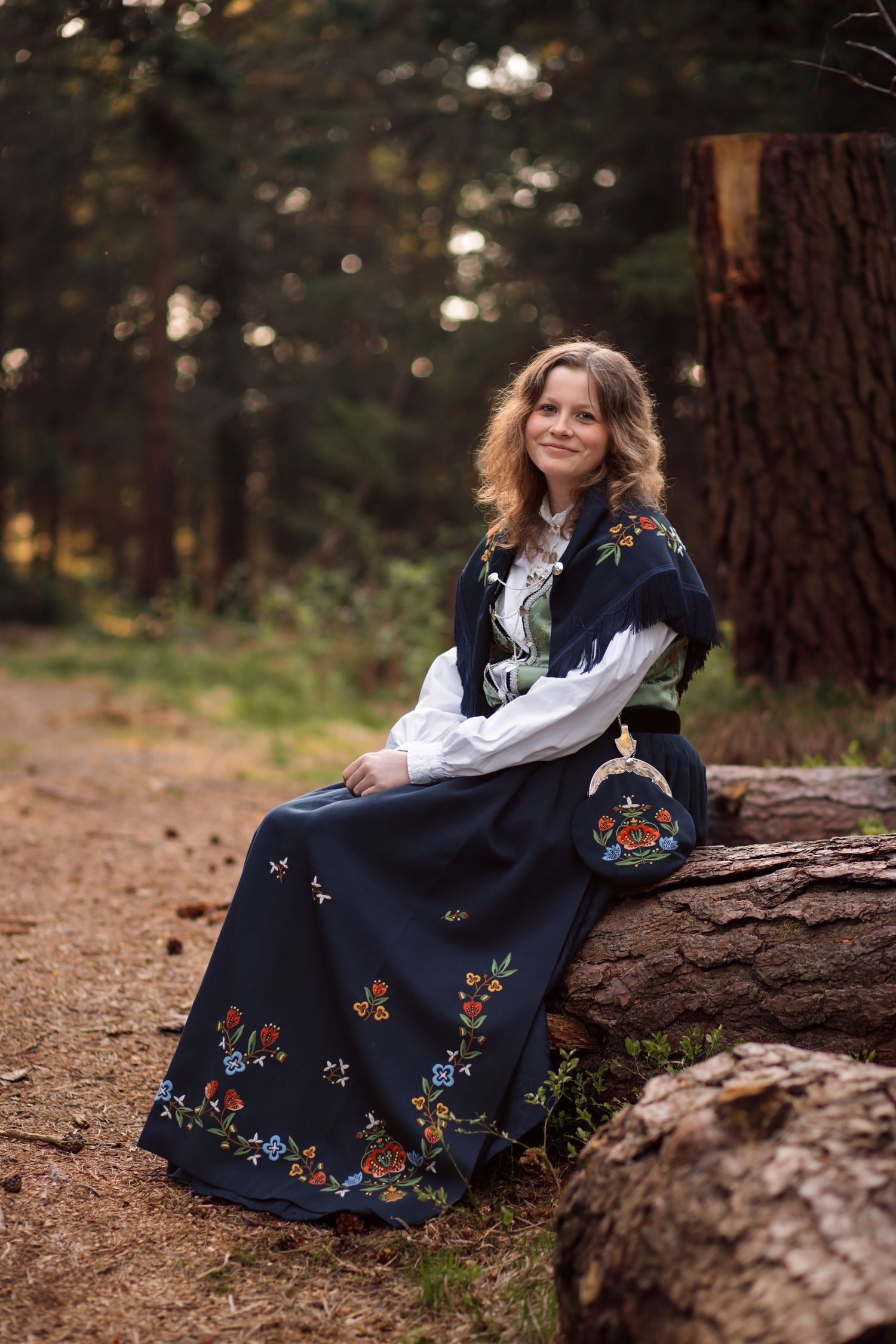A person in a white blouse and navy floral dress sits on a fallen log in a forest setting with dappled sunlight.