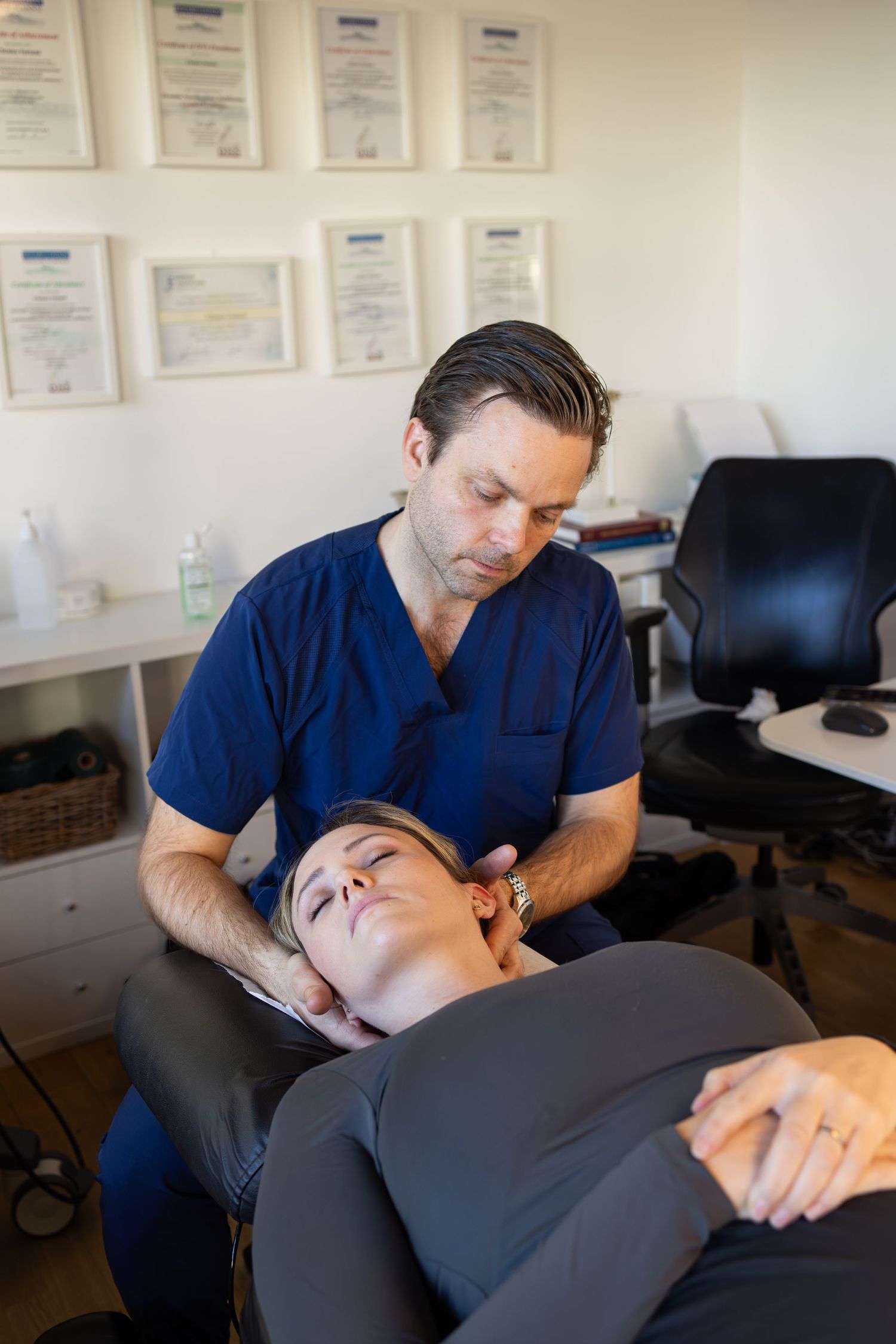 A chiropractor in blue scrubs performs neck adjustment therapy on a patient lying on a treatment table.