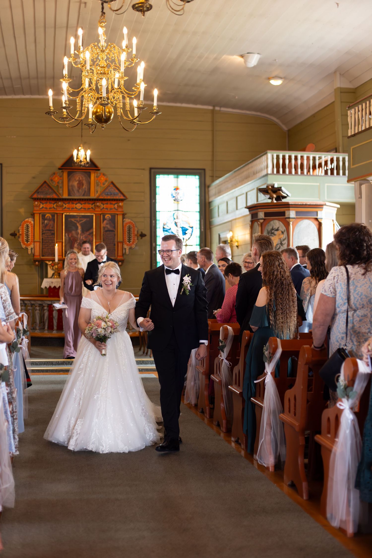 A couple walks down the aisle of a historic church with wooden pews and elegant chandeliers during a wedding ceremony.