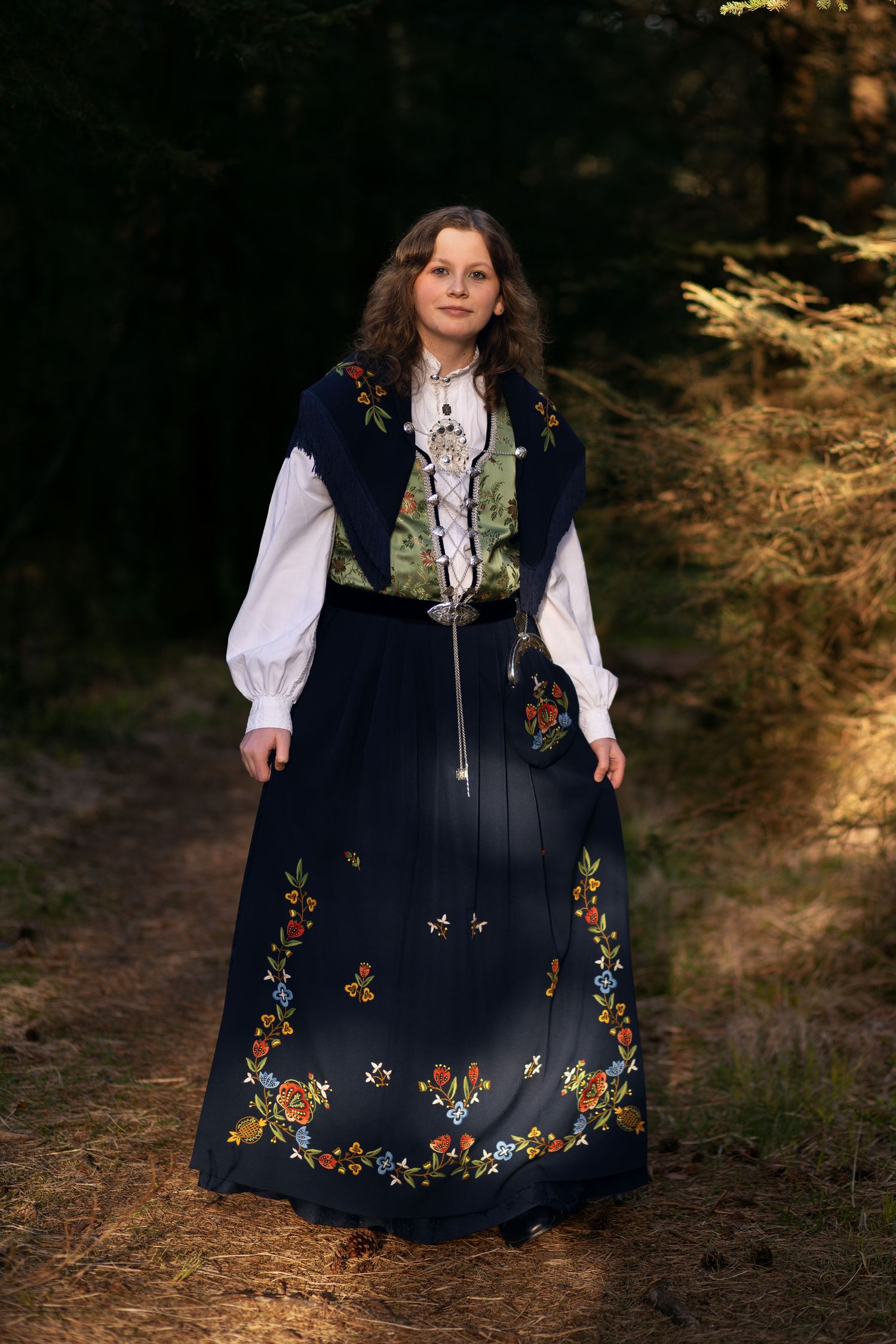 A person wearing a traditional embroidered navy blue dress with floral patterns stands in a sunlit forest setting.