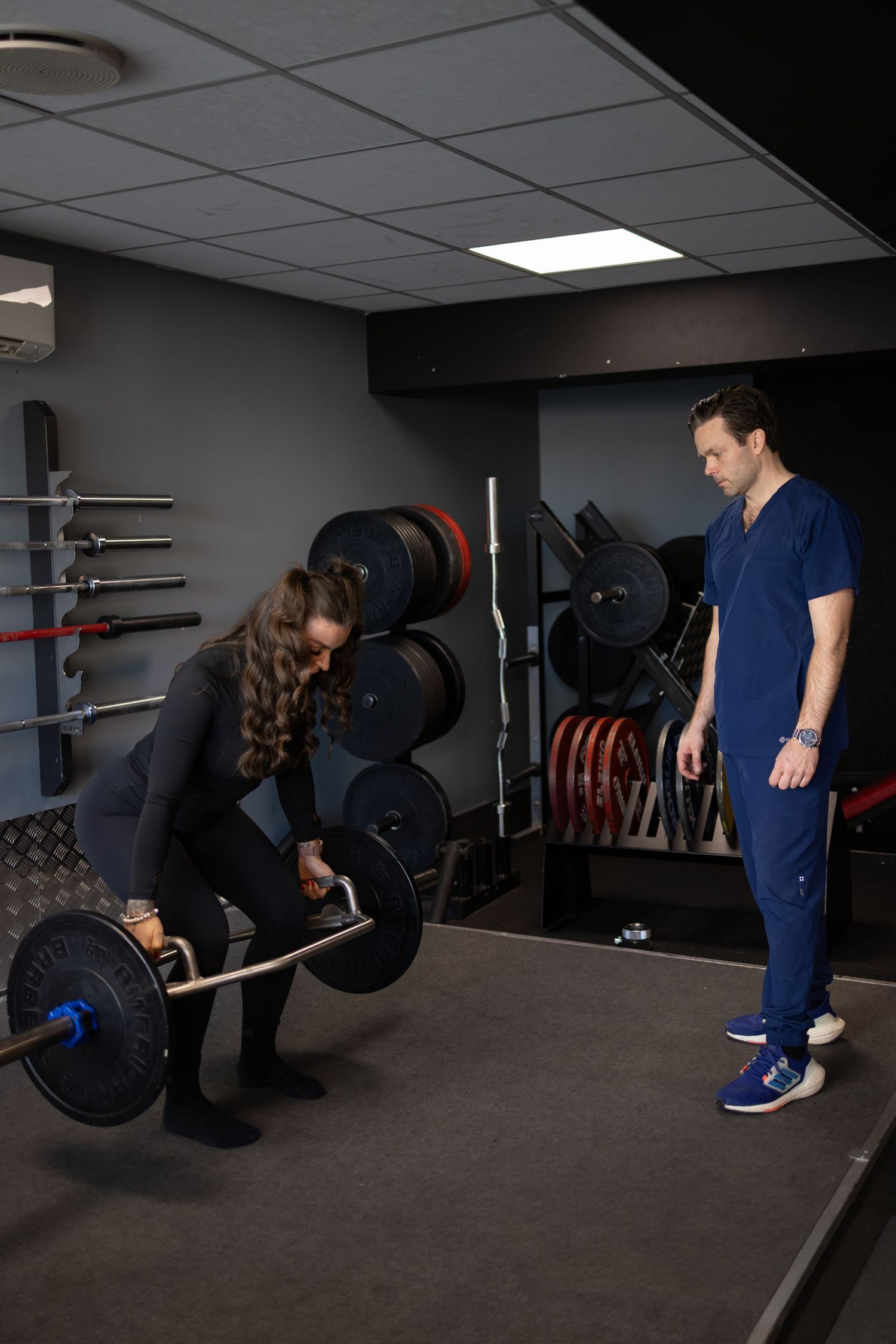 Two people in a gym setting with weight plates and equipment, one preparing to lift a barbell while the other observes. personlig trener