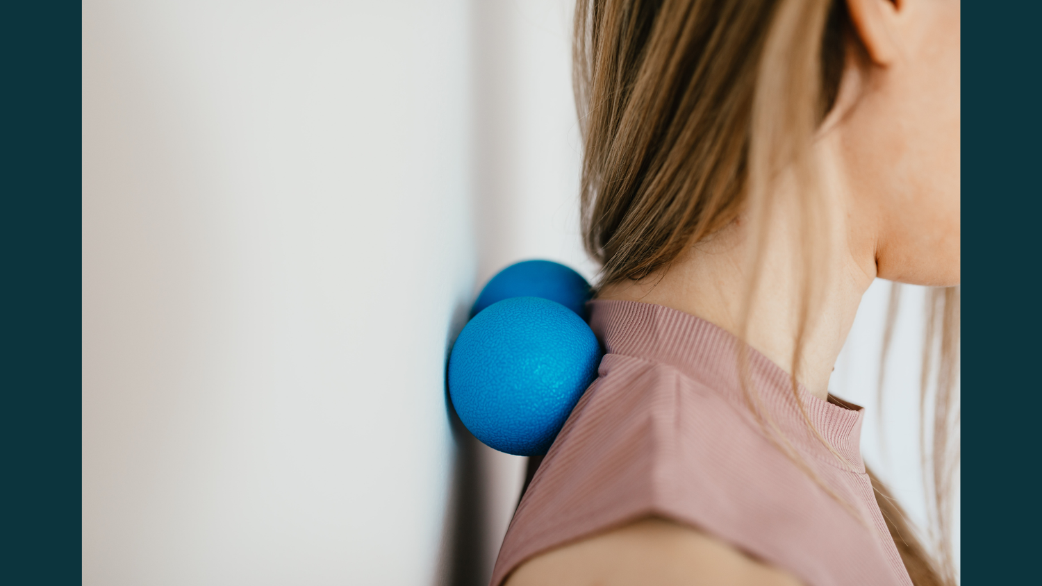 A woman using blue massage therapy balls against a wall to apply pressure to her upper back and neck for self-myofascial release.