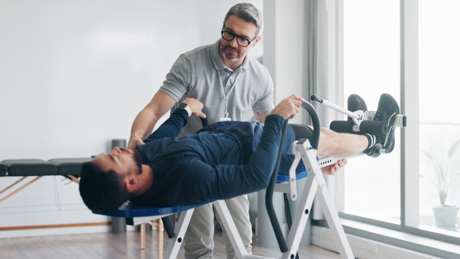 A male therapist in glasses assisting a patient on a spinal decompression table, adjusting the equipment for a rehabilitation session.