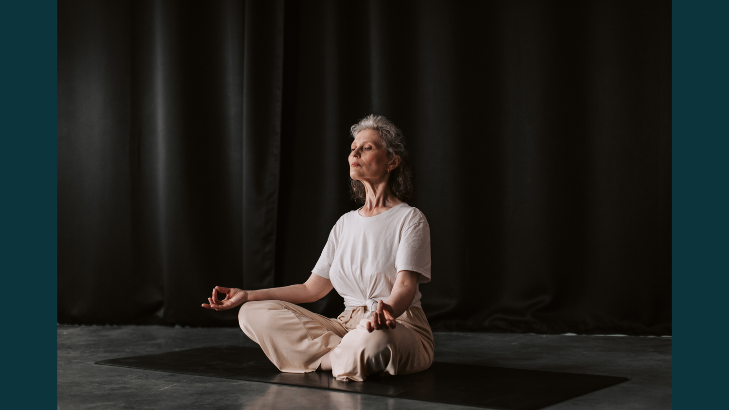 An older woman with grey hair sits in a meditative yoga pose on a black mat, wearing a white t-shirt and beige trousers against dark curtain