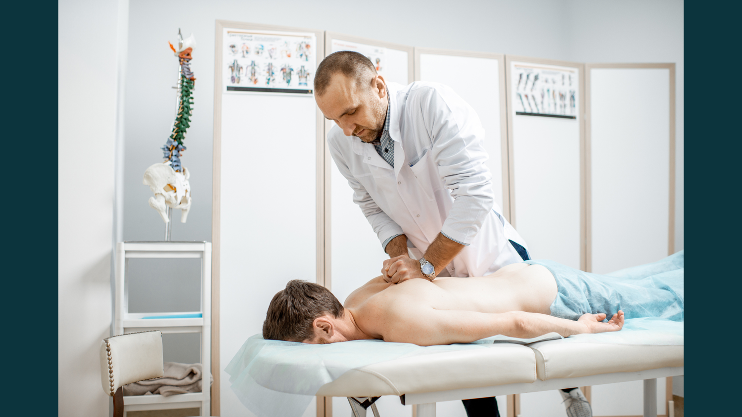 A male practitioner in a white coat performing manual spinal mobilisation on a patient lying prone on a treatment table.