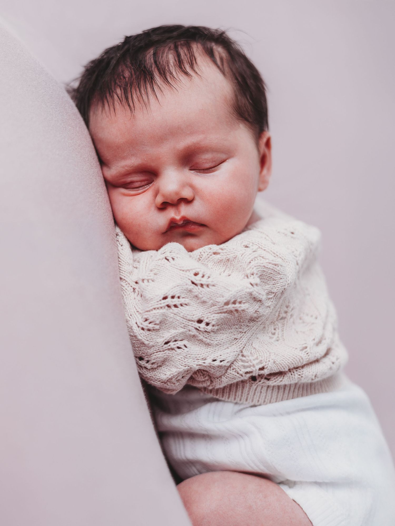 A sleeping baby wrapped in a delicate white lace blanket rests peacefully against a soft pink background.