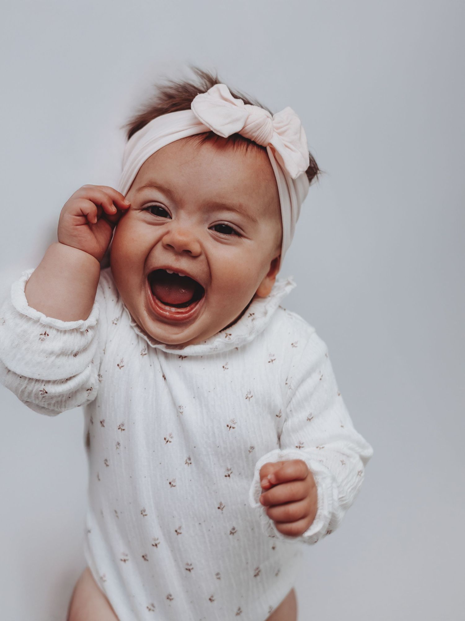 A laughing baby girl wearing a white floral onesie and pink bow headband against a light gray background.